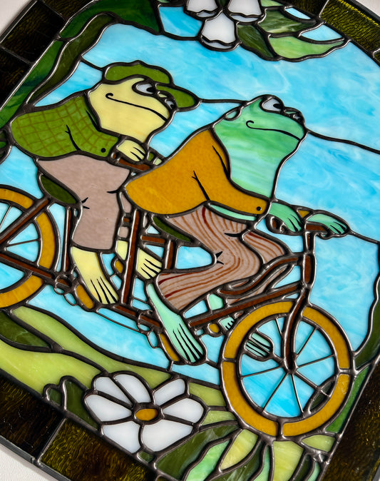 close up of a stained glass panel depicting frog and toad riding a bicycle resting on a white surface