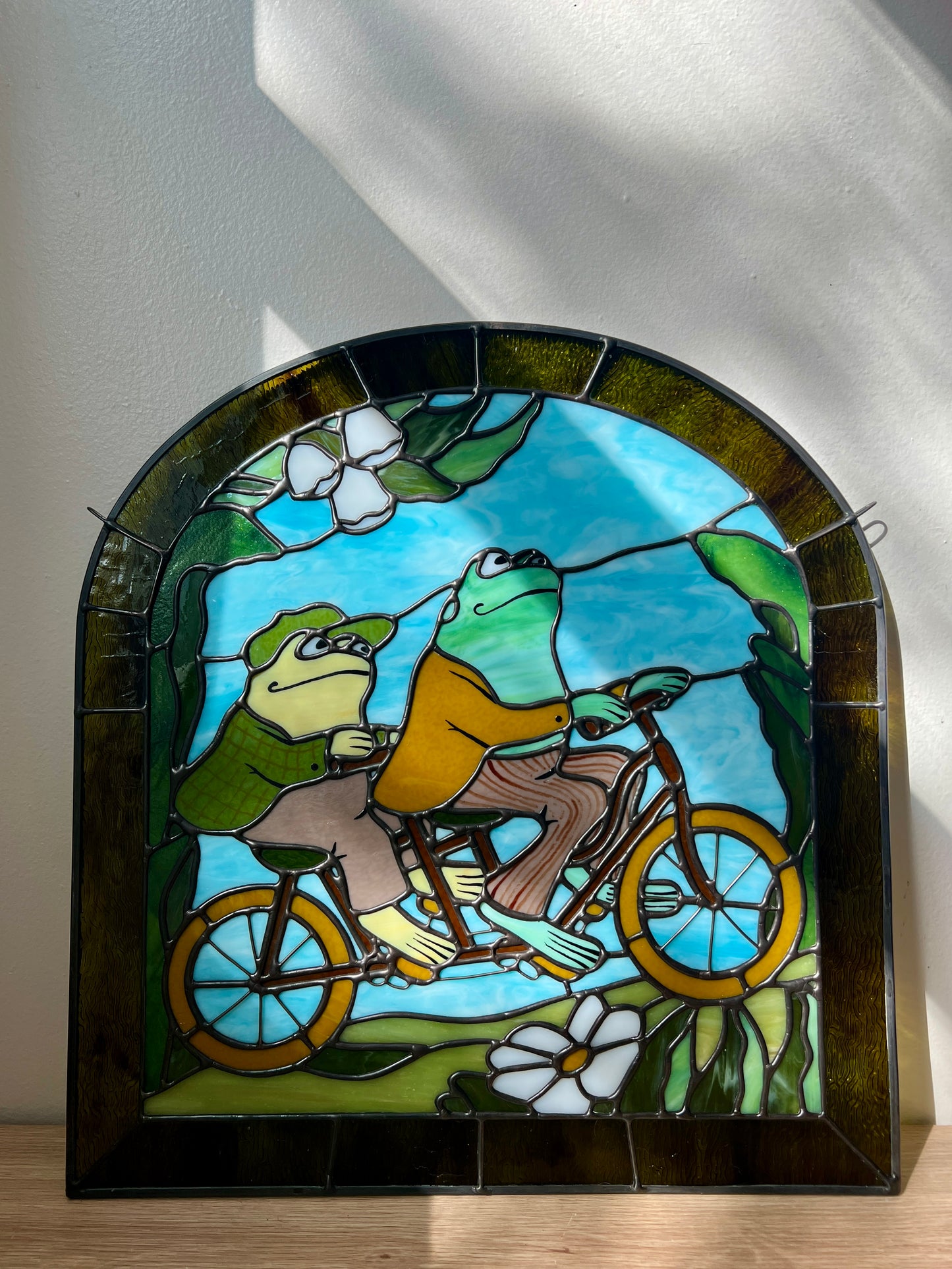stained glass panel depicting frog and toad riding a bicycle resting on a table leaning against a white wall