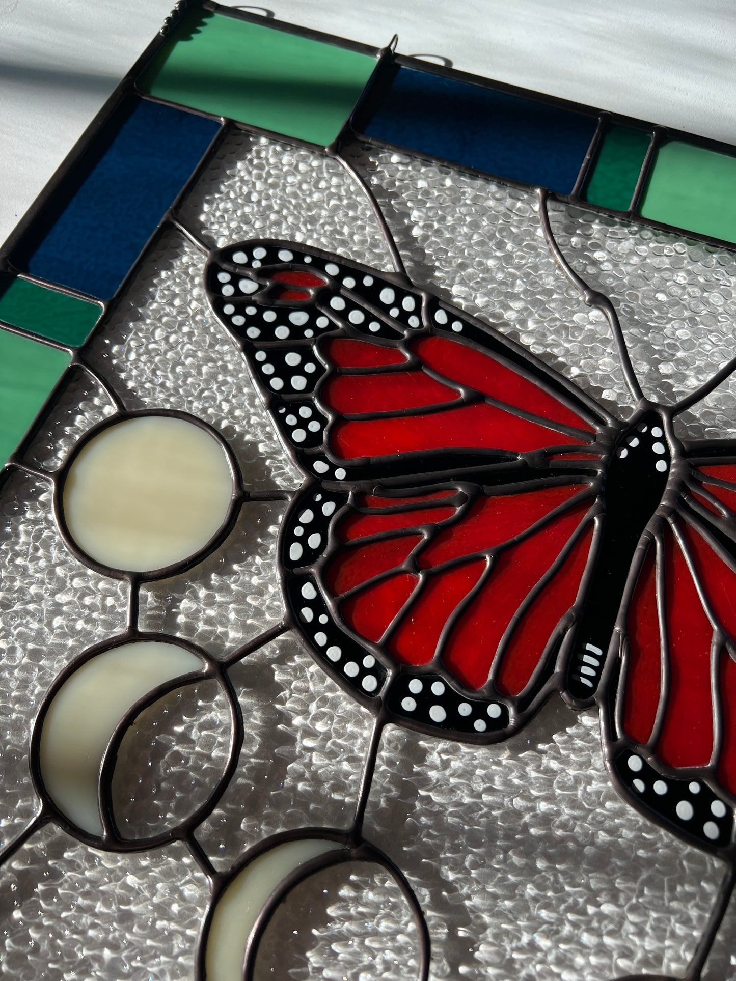 A large stained glass panel with a thick blue and green geometric border and a monarch butterfly and moon phases in the center, with textured clear glass in the background. The image provides a close up of the left half of the design, and is photographed at an angle.