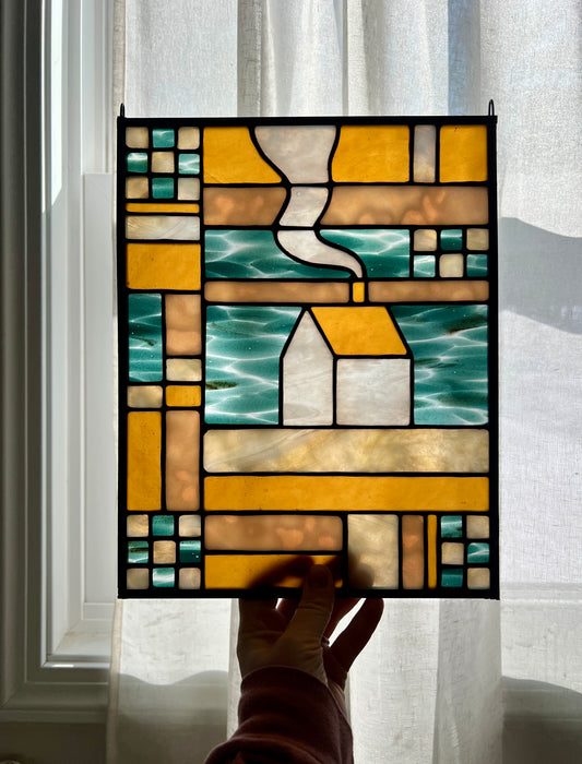 Stained glass textile inspired panel depicting a home in the center in shades of teal, amber, white, and beige, held by a hand against a brightly backlit sheer white curtain