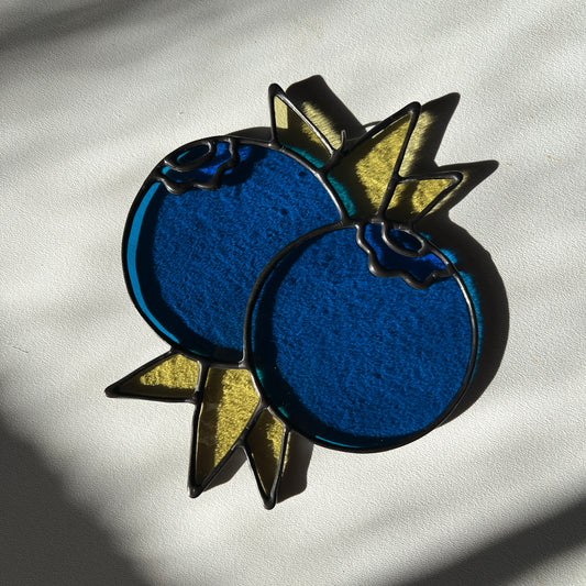 a stained glass suncatcher depicting a small bunch of blueberries with leaves resting on a white surface