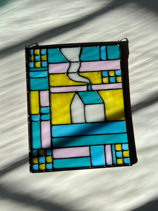 Stained glass textile inspired panel depicting a home in the center in shades of teal, pink, white, blue, and yellow, resting against a white background white bright sunlight and shadows flowing across it