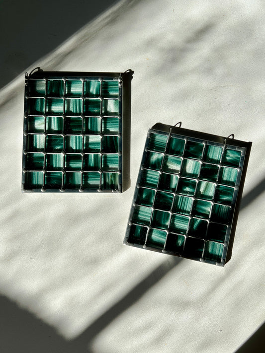 two stained glass suncatchers depicting streaky and mottled sparkly clear and teal glass arranged in a grid pattern, resting on a white surface