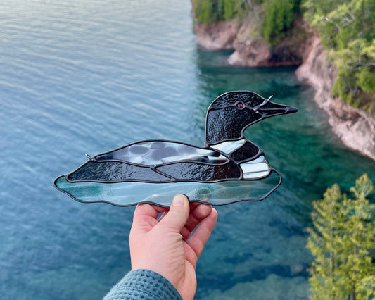 Hand holding a stained glass loon ornament over a cliffside overlooking turquoise colored lake superior and opposing cliff shoreline.
