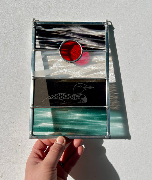 a hand holding a stained glass panel with black, white, clear, teal, and red glass with a loon etched on the surface