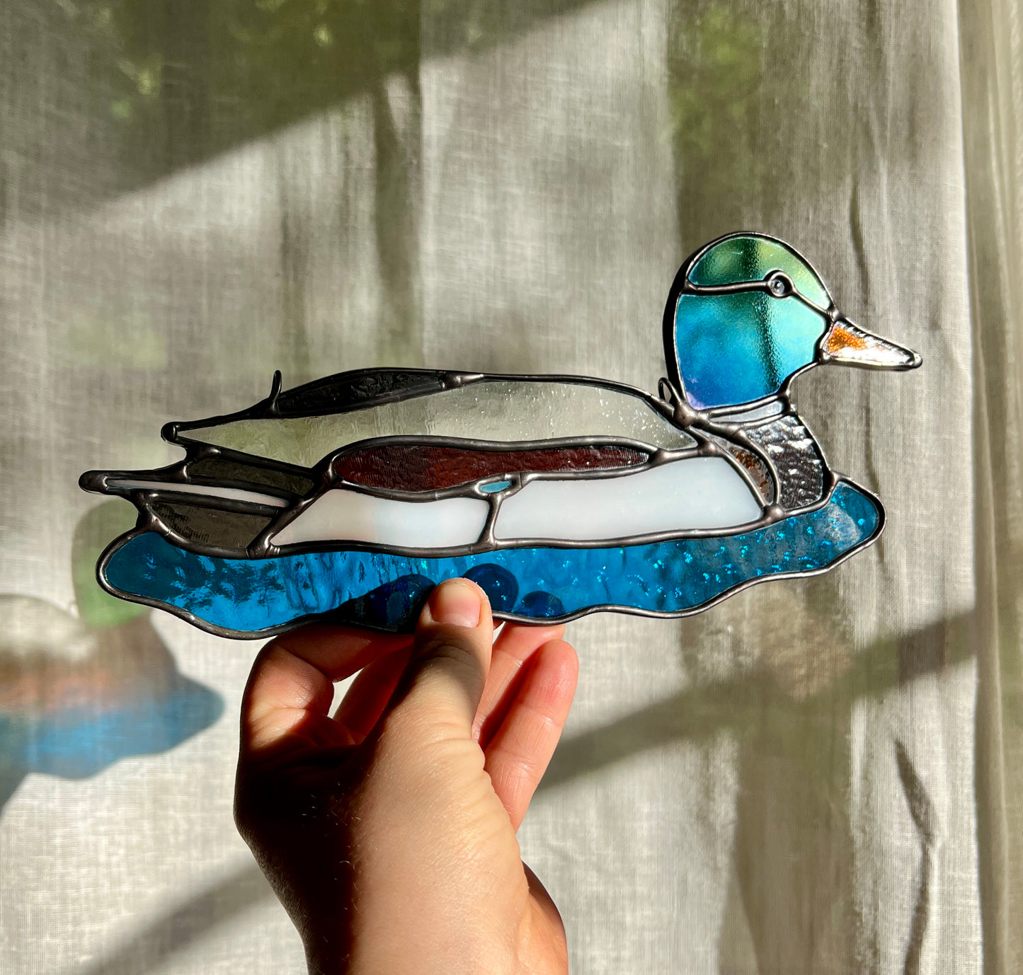 stained glass suncatcher of a male mallard held by a hand at an angle to demonstrate green and blue iridescence of the glass used for the head