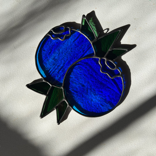 a wispy blue stained glass suncatcher depicting a small bunch of blueberries with leaves resting on a white surface
