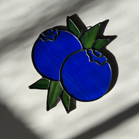 stained glass suncatcher depicting a small bunch of blueberries with leaves resting on a white surface