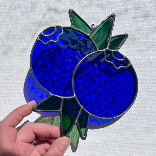 A hand holding a stained glass suncatcher depicting two blueberries and some leaves at an angle against a white background.