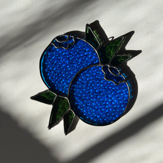 a textured stained glass suncatcher depicting a small bunch of blueberries with leaves on a white surface