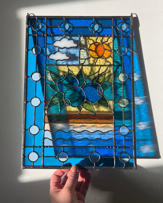 stained glass panel with a simple compartment style design featuring clouds, sun, water, and a small bunch of blueberries in the middle with a blue border held by a hand against a wall