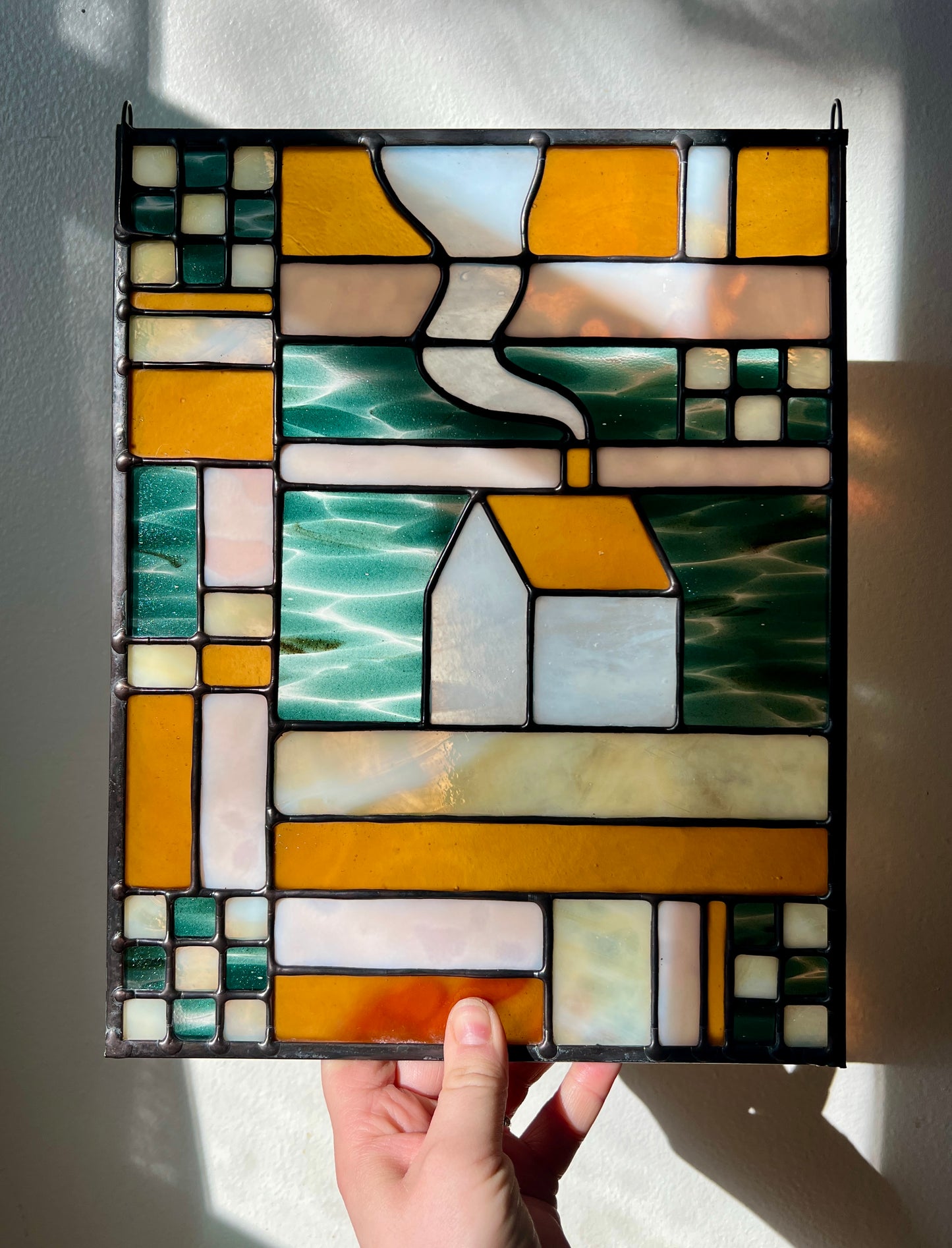 Stained glass textile inspired panel depicting a home in the center in shades of teal, amber, white, and beige, held by a hand against a white wall with bright sunlight cast across