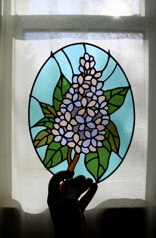 Stained glass panel with floral lilac design held in front of a window with sheer curtains.