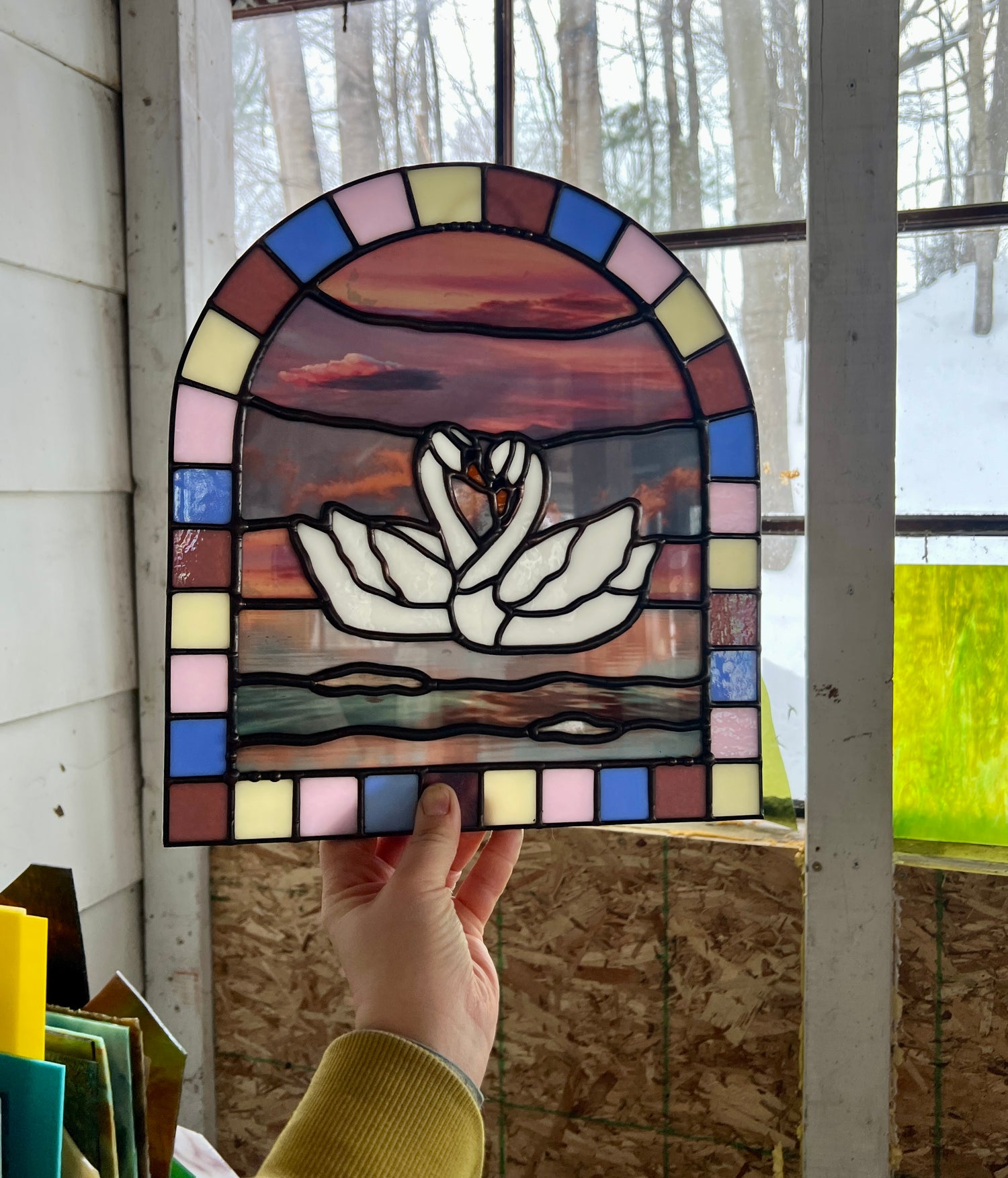 Stained glass panel with a colorful pastel arch border and two white swans in the center. Collaged photographs are used to create a sunset background. The art piece is held up by a hand indoors.