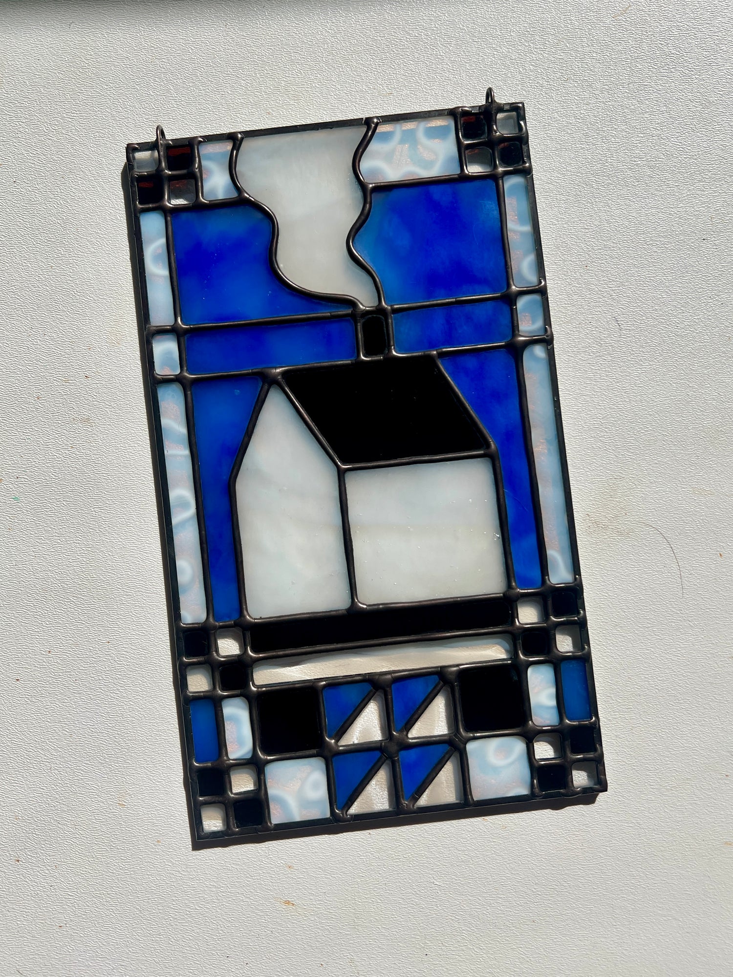 Stained glass textile inspired panel depicting a home in the center in shades of blue, brown, and white, resting on a white surface