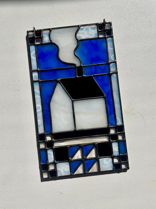 Stained glass textile inspired panel depicting a home in the center in shades of blue, brown, and white, resting on a white surface