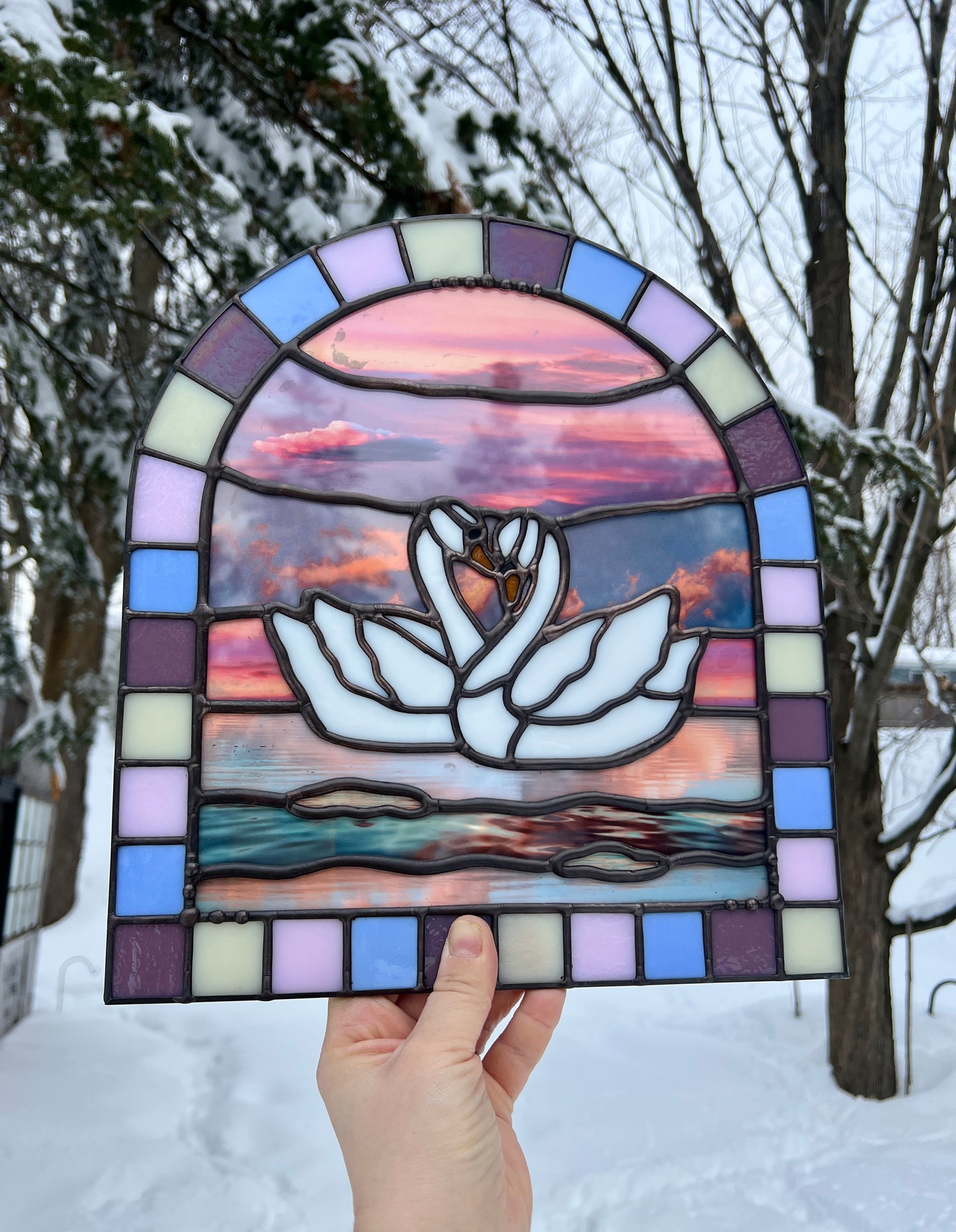 Stained glass panel with a colorful pastel arch border and two white swans in the center. Collaged photographs are used to create a sunset background. The piece is held by a hand with a winter landscape in the background.