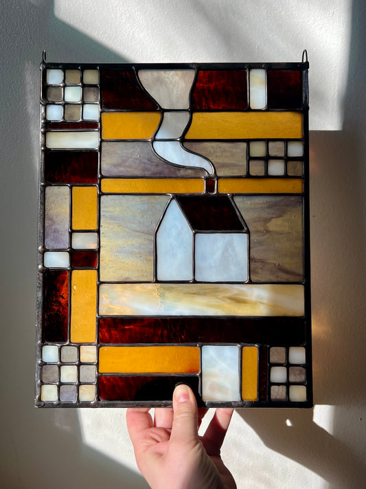 Stained glass textile inspired panel depicting a home in the center in shades of purple, white, brown, and amber, held by a hand in front of a white surface