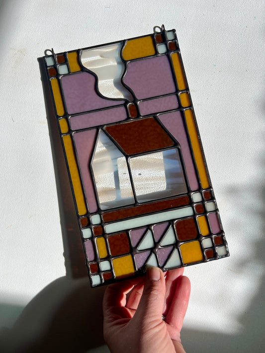 Stained glass textile inspired panel depicting a home in the center in shades of purple, white, brown, and bronze, held by a hand against a white surface with bright sunlight cast across.