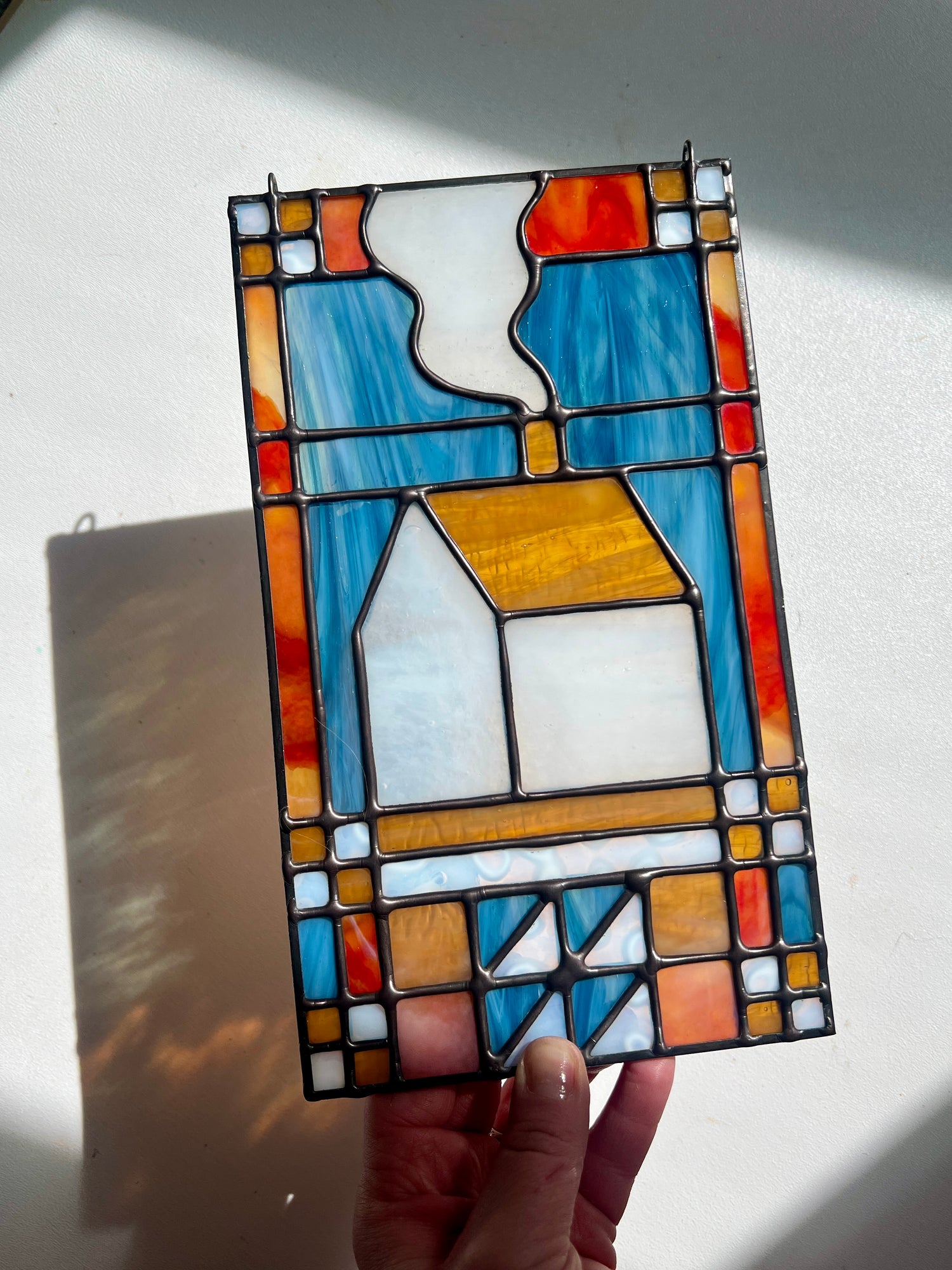 Stained glass textile inspired panel depicting a home in the center in shades of red, blue, white, and amber, held by a hand in front of a white background
