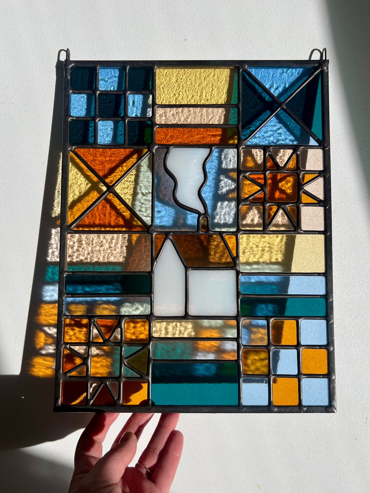 Stained glass textile inspired panel depicting a home in the center in shades of teal, pink, white, blue, and yellow, held by a hand against a white background with bright sunlight casting subtle refractions