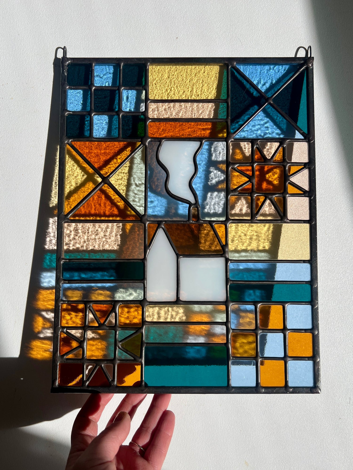 Stained glass textile inspired panel depicting a home in the center in shades of teal, pink, white, blue, and yellow, held by a hand against a white background with bright sunlight casting subtle refractions