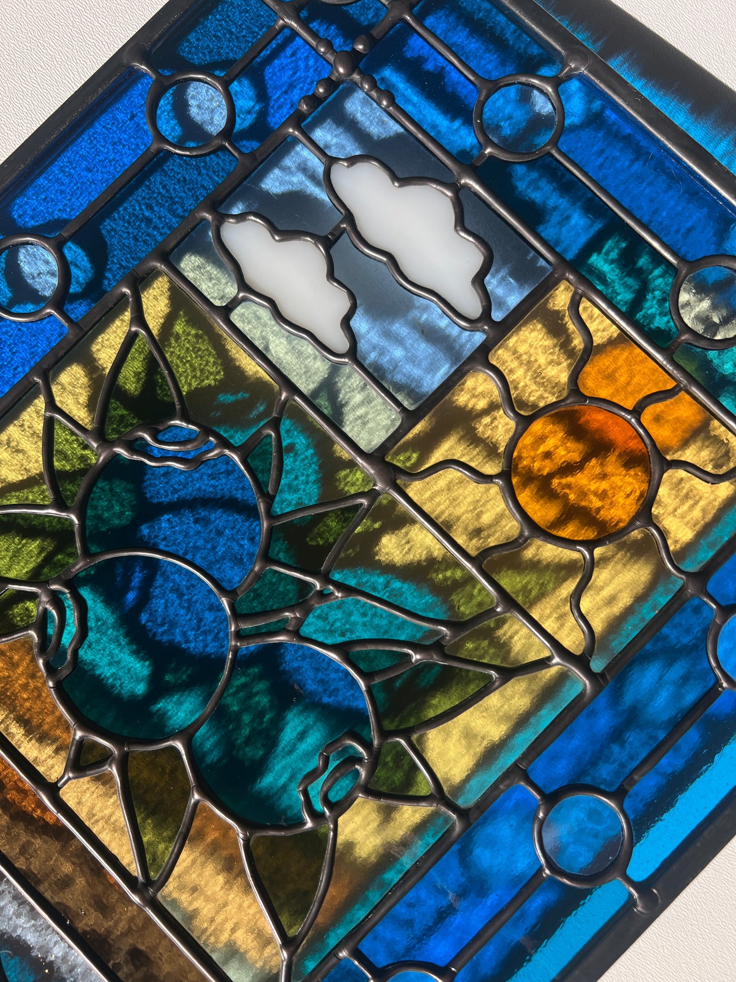 close up of a stained glass panel with a simple compartment style design featuring clouds, sun, water, and a small bunch of blueberries in the middle with a blue border