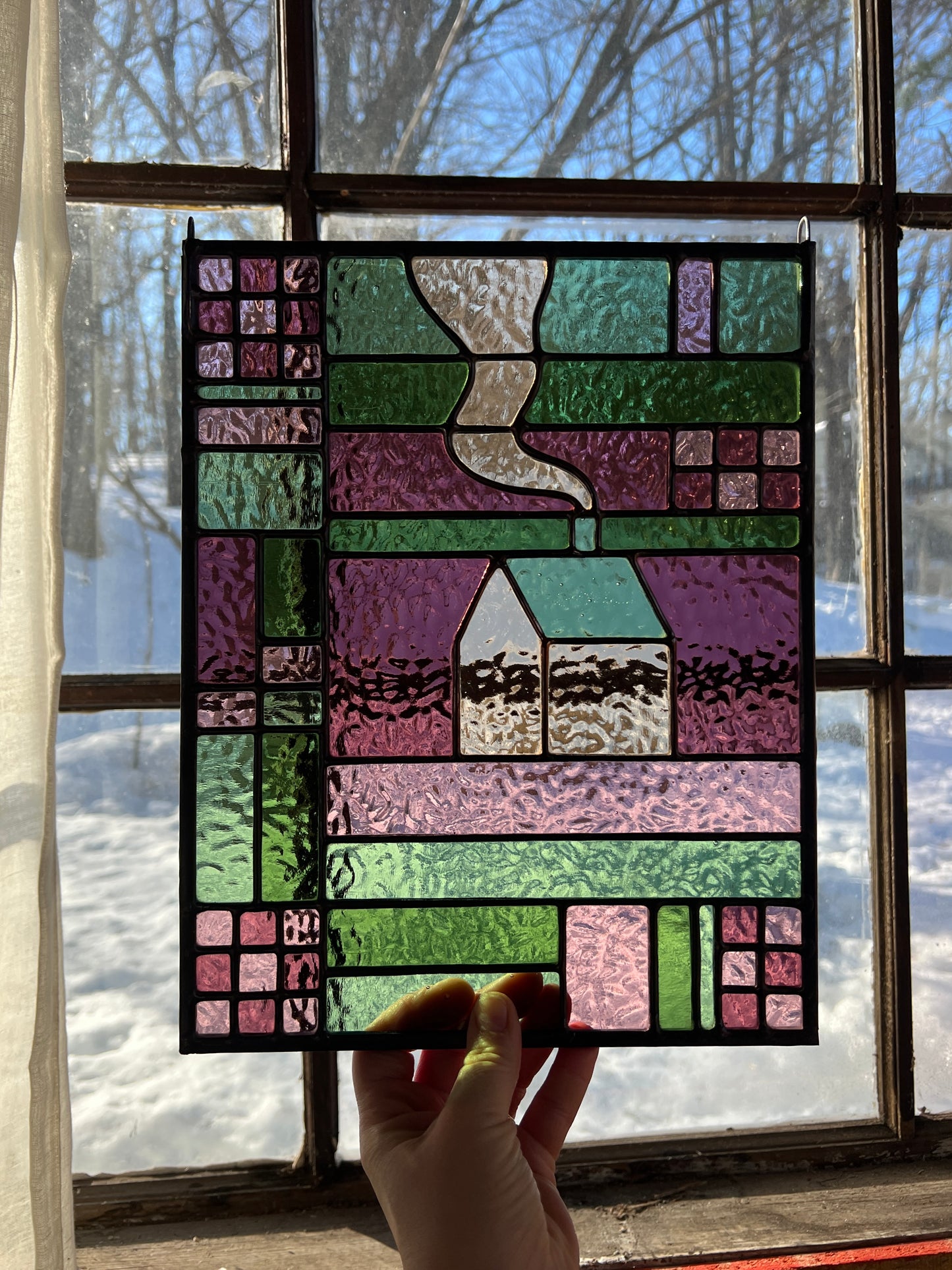 Hand holding a stained glass panel with a house design in front of a window with a snowy landscape.