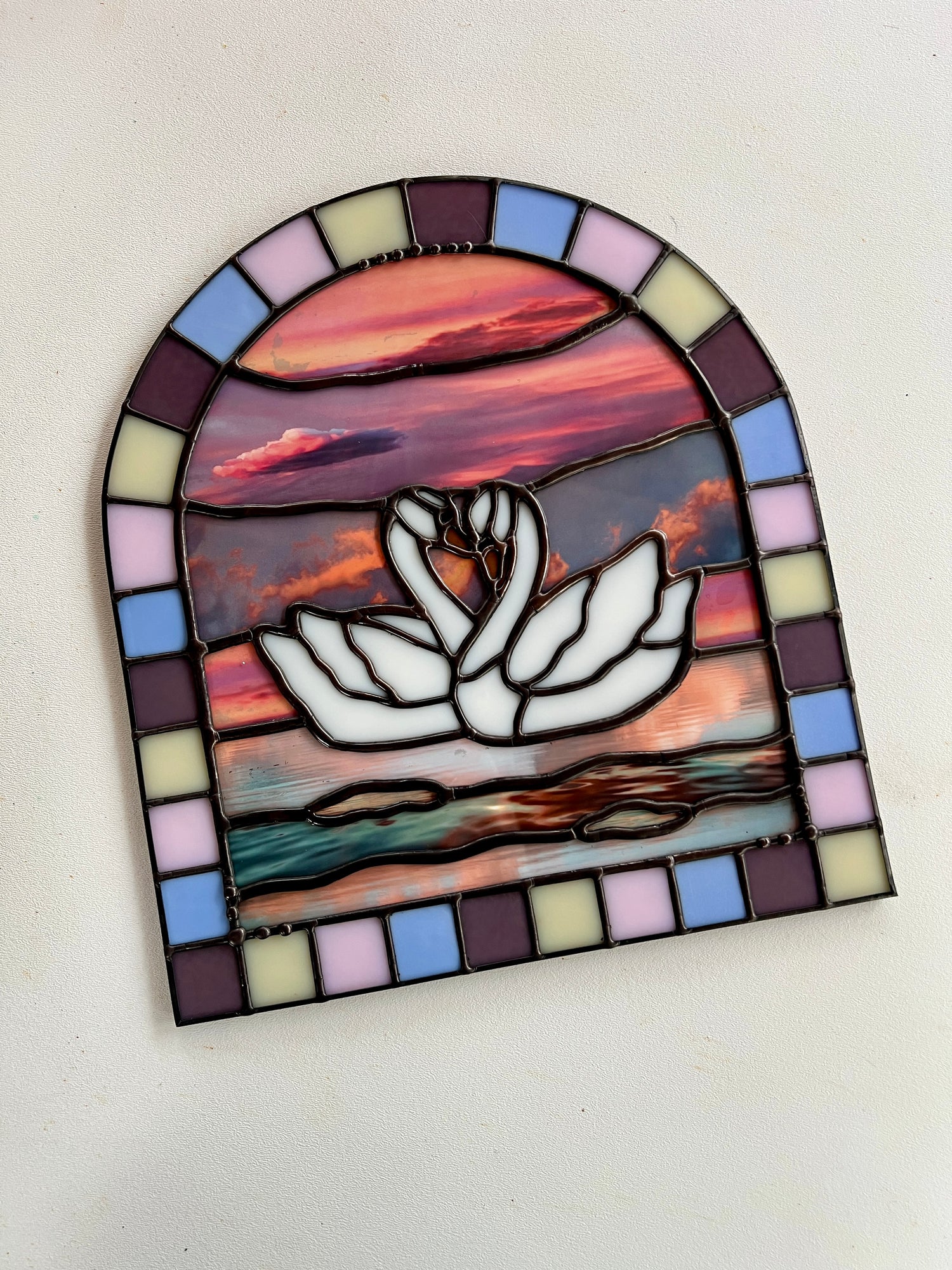 Stained glass panel with a colorful pastel arch border and two white swans in the center. Collaged photographs are used to create a sunset background.