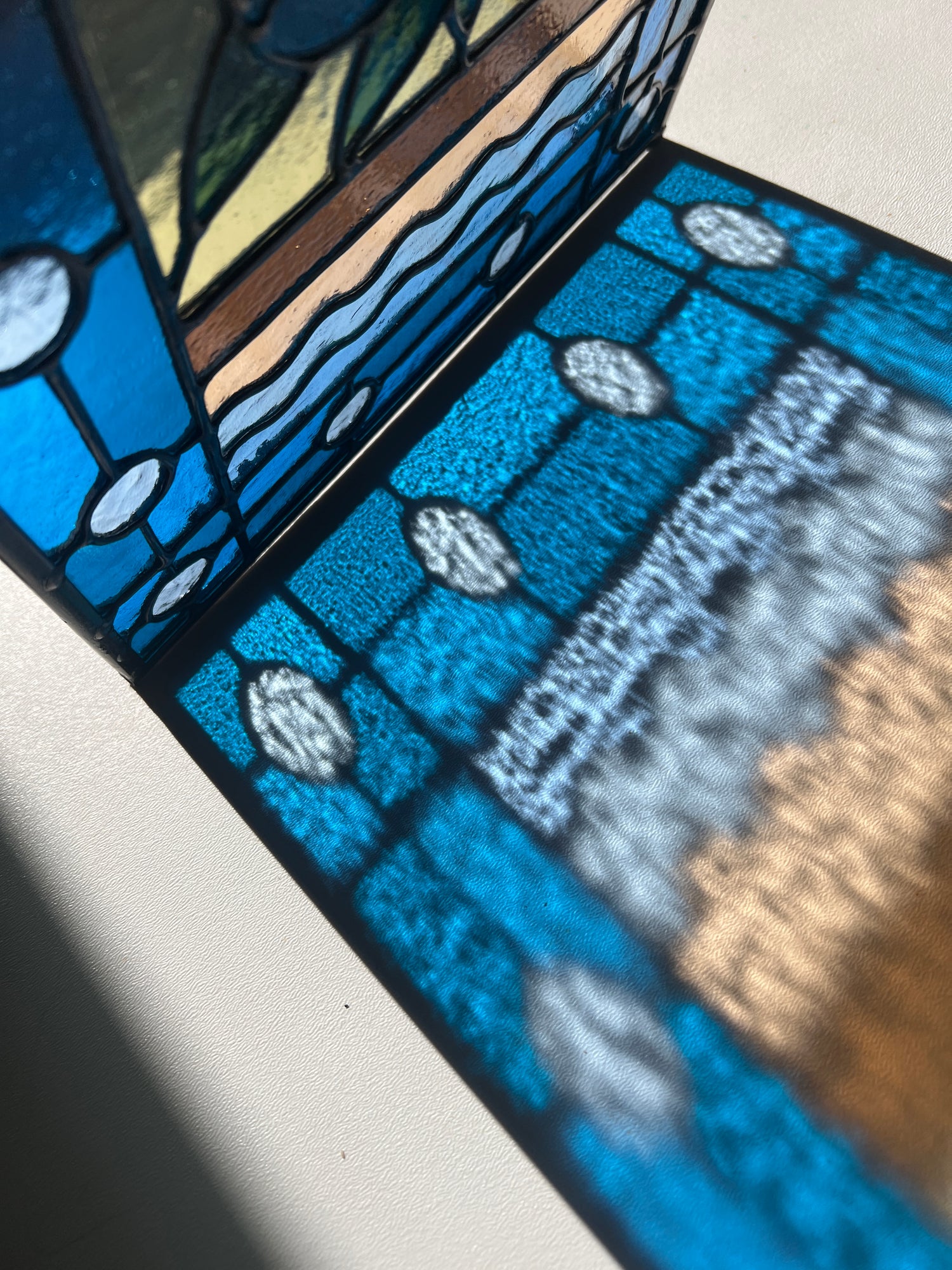 close up of a refraction of a stained glass panel with a simple compartment style design featuring clouds, sun, water, and a small bunch of blueberries in the middle with a blue border