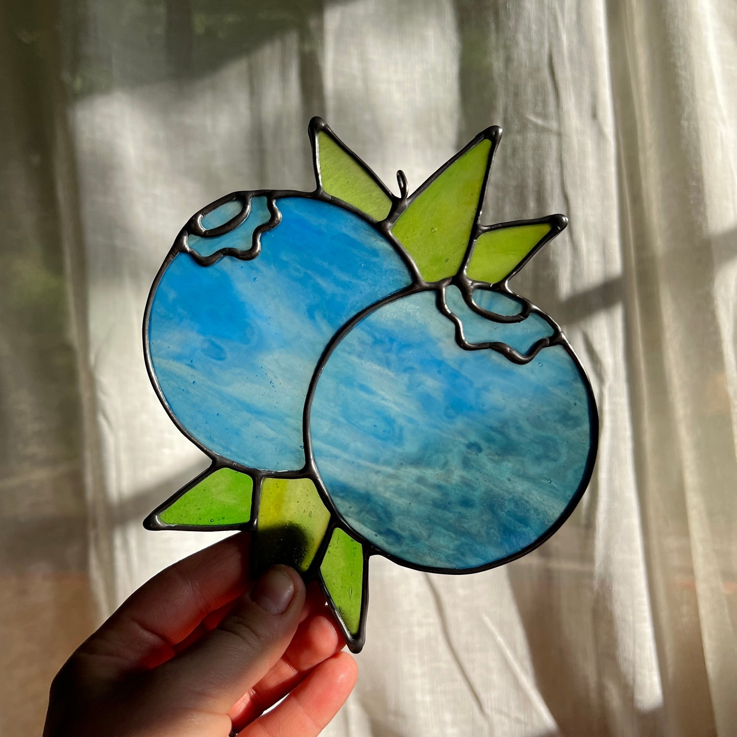 hand holding a  wispy light blue stained glass suncatcher depicting a small bunch of blueberries with leaves in front of a window