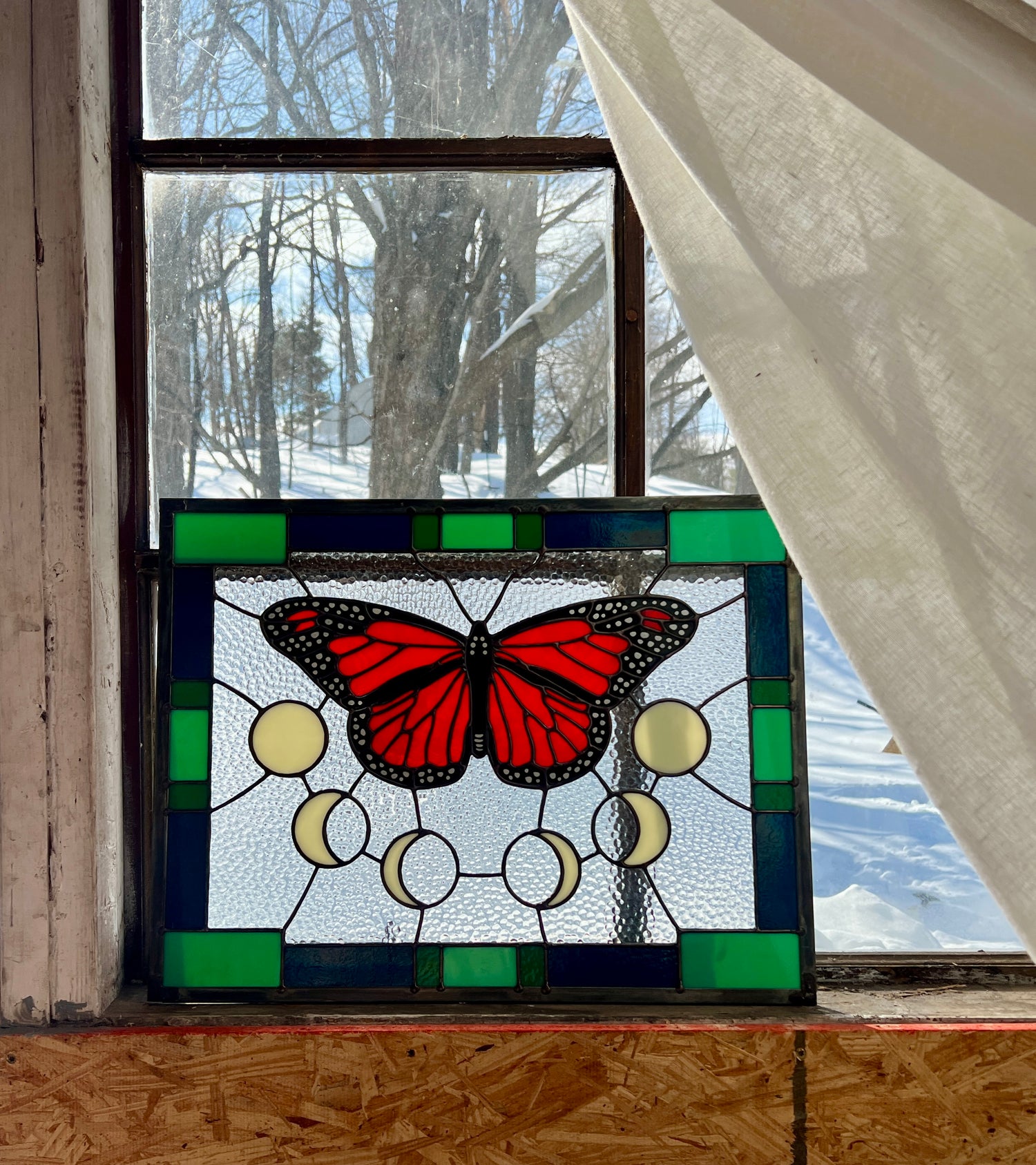 A large stained glass panel with a thick blue and green geometric border and a monarch butterfly and moon phases in the center, with textured clear glass in the background, resting against a window with a snowy landscape outside and a sheer white curtain pulled aside.