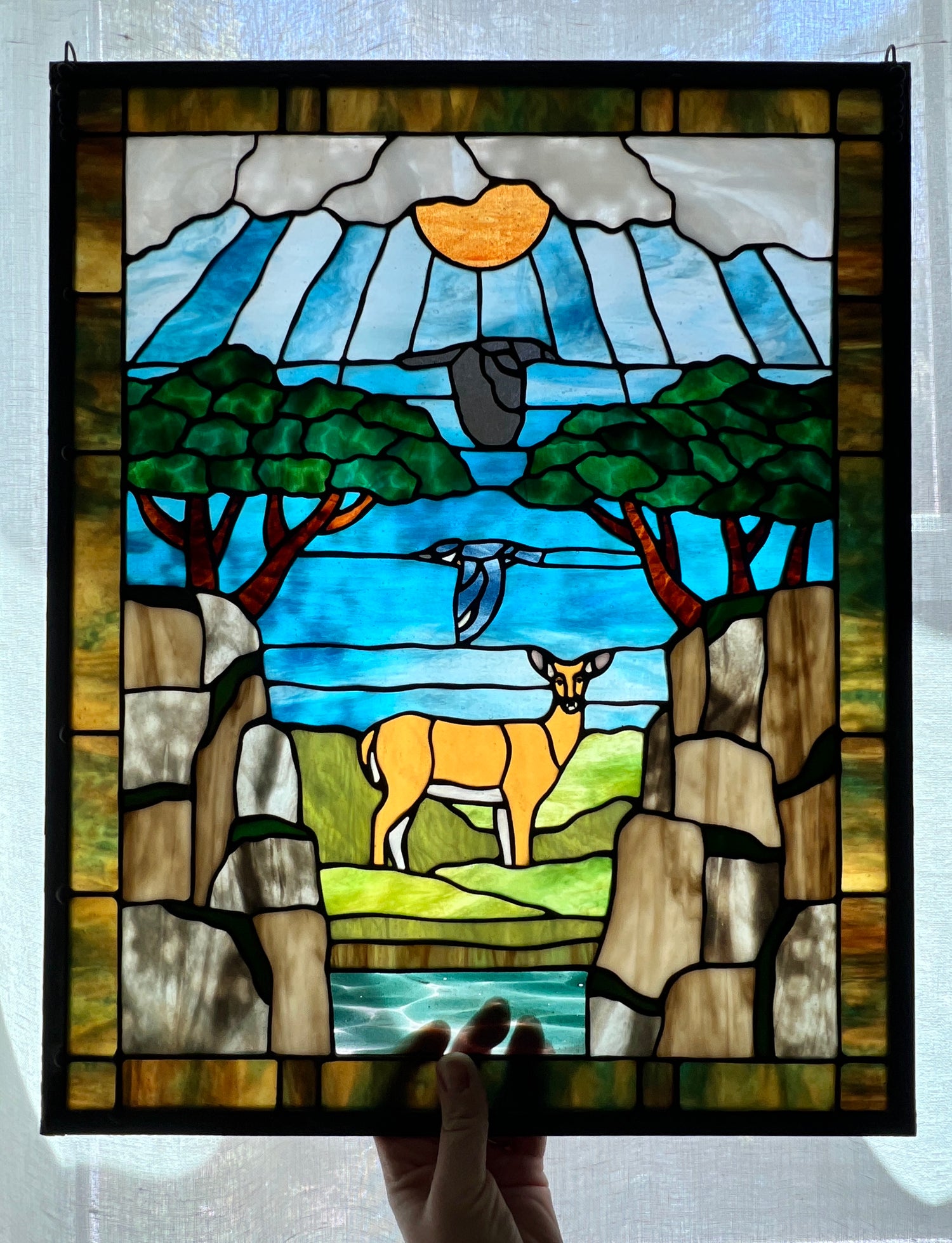 large stained glass panel depicting mossy, rocky cliffs and trees, with a deer, a kingfisher, and a raven standing amongst a river, greenery, and bright blue skies with some clouds and a bright yellow orange sun.