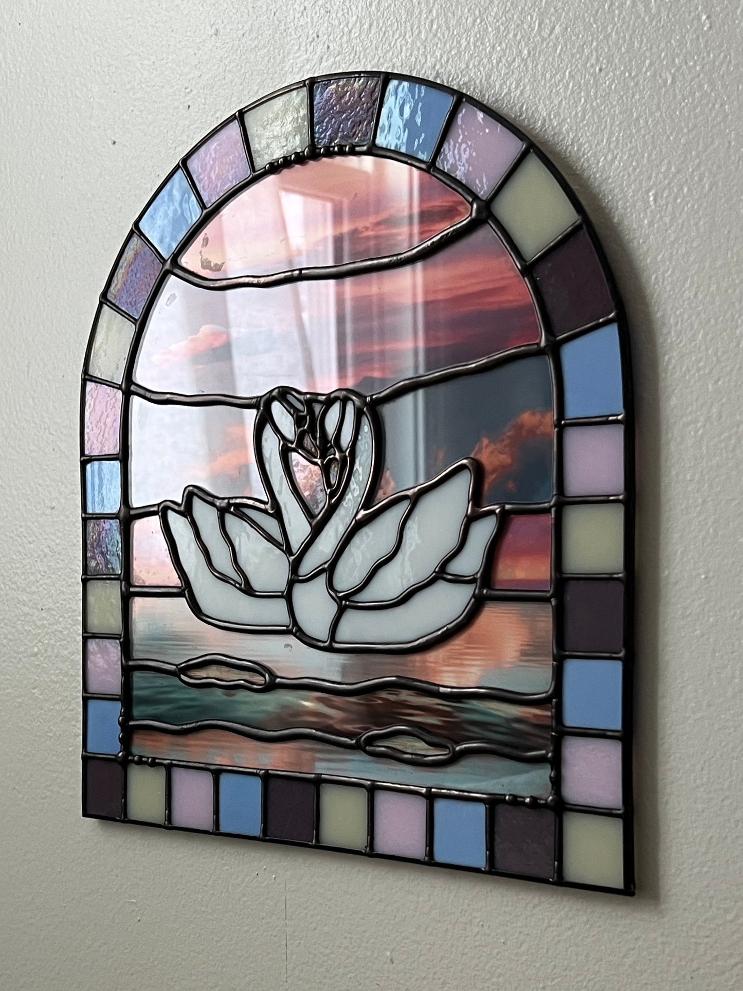 Stained glass panel with a colorful pastel arch border and two white swans in the center. Collaged photographs are used to create a sunset background. The piece is hanging up against a white wall and photographed at an angle.