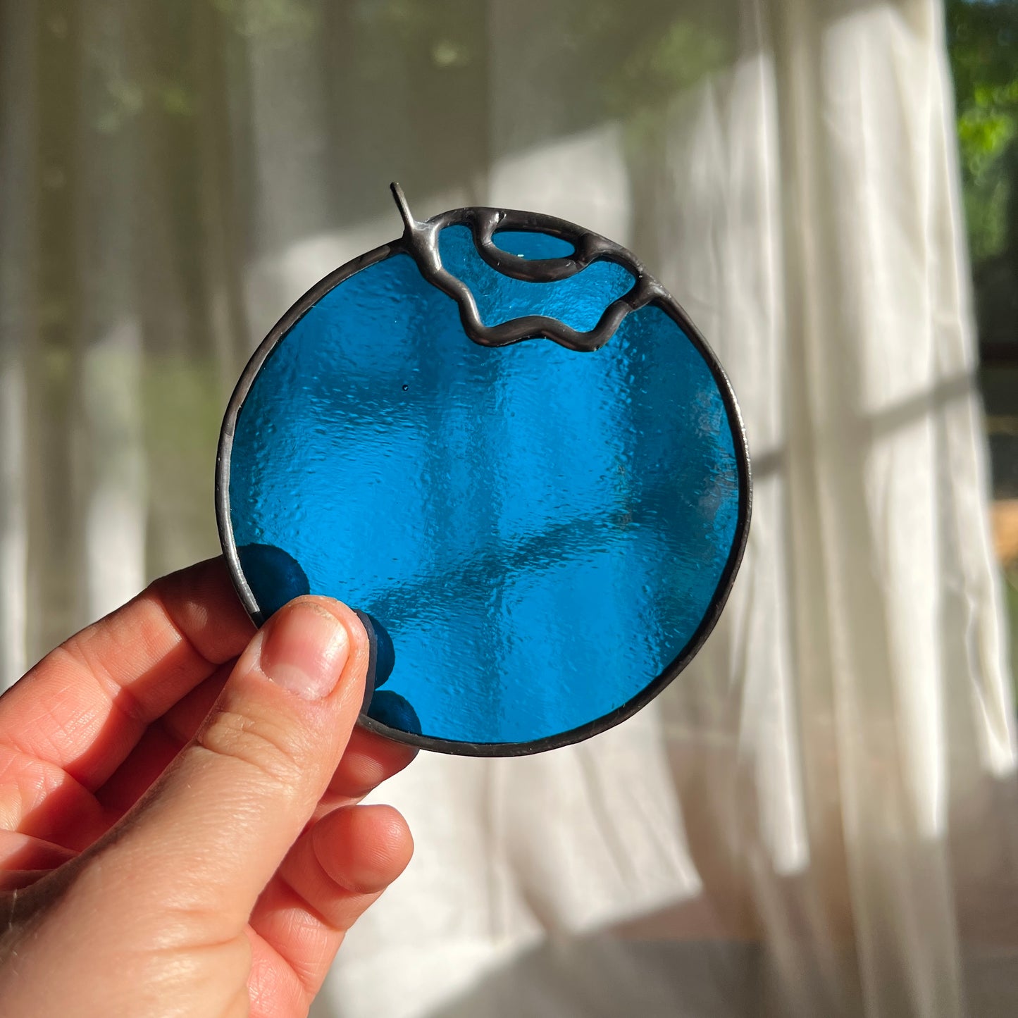 hand holding a wispy blue stained glass blueberry in front of a window