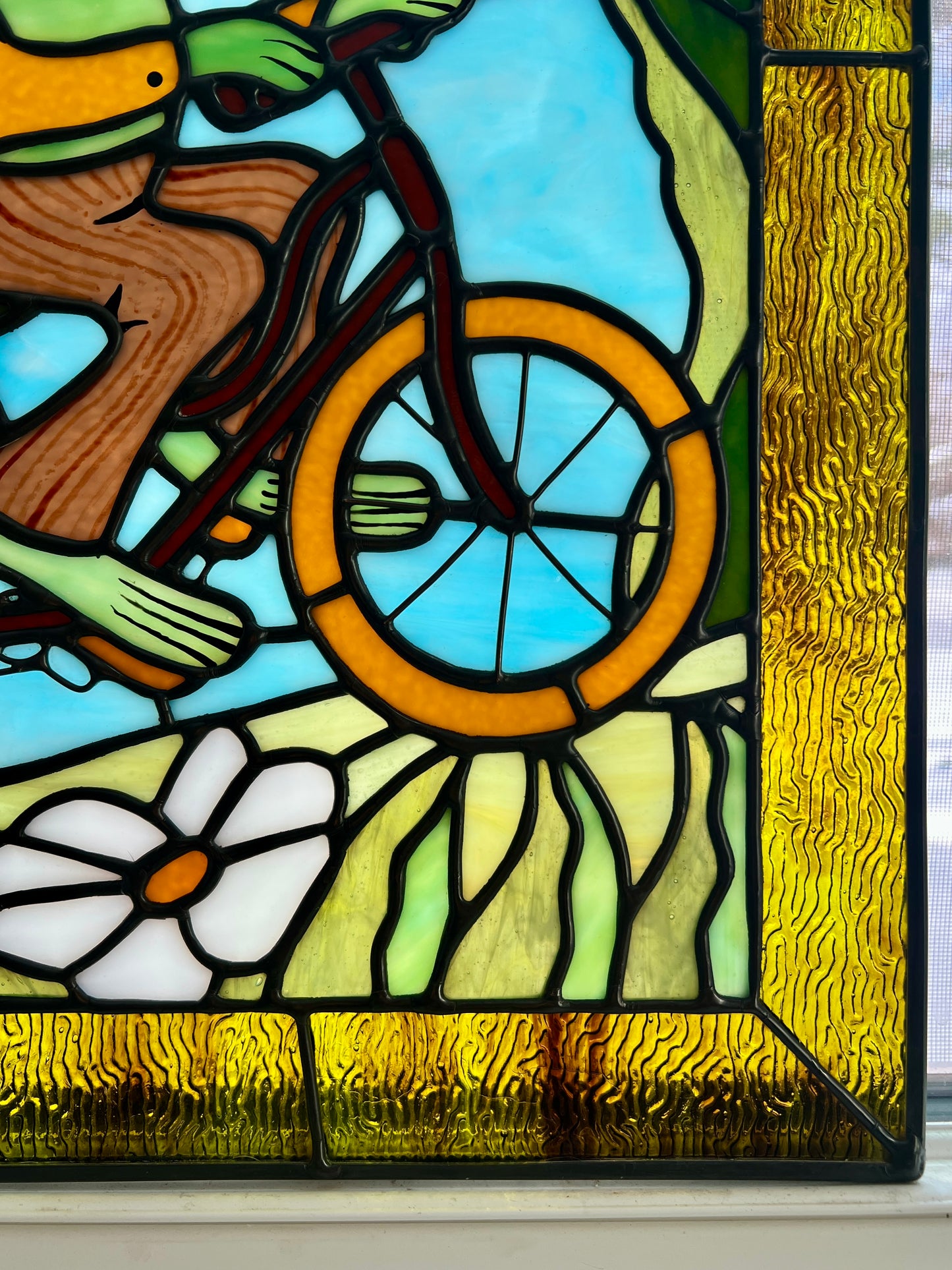 close up of a stained glass panel depicting frog and toad riding a bicycle with soft backlighting
