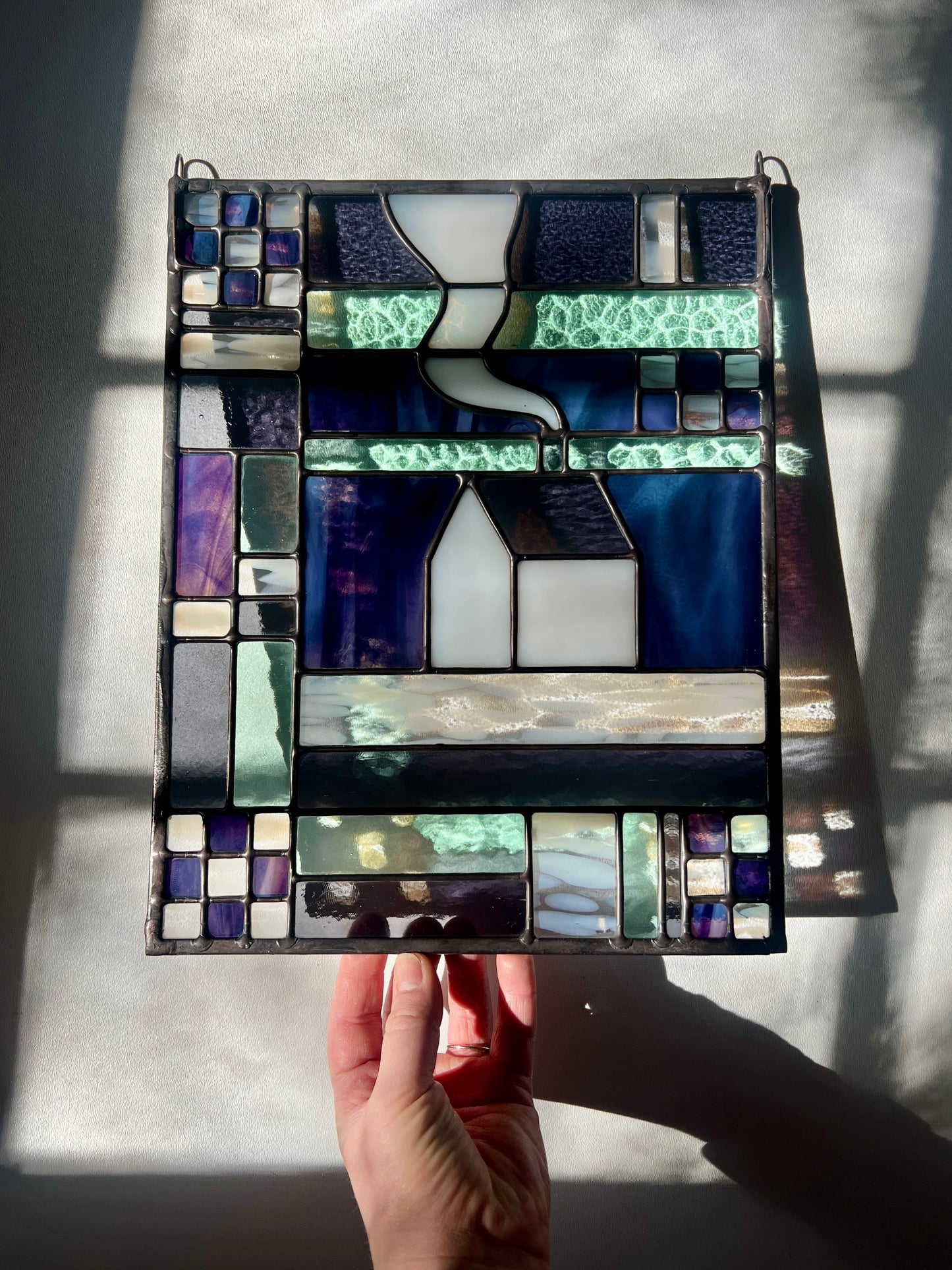 Stained glass panel with geometric textile inspired design with a cozy home depicted in the center, with a hand holding it against a white background allowing subtle colorful refractions from the stained glass.
