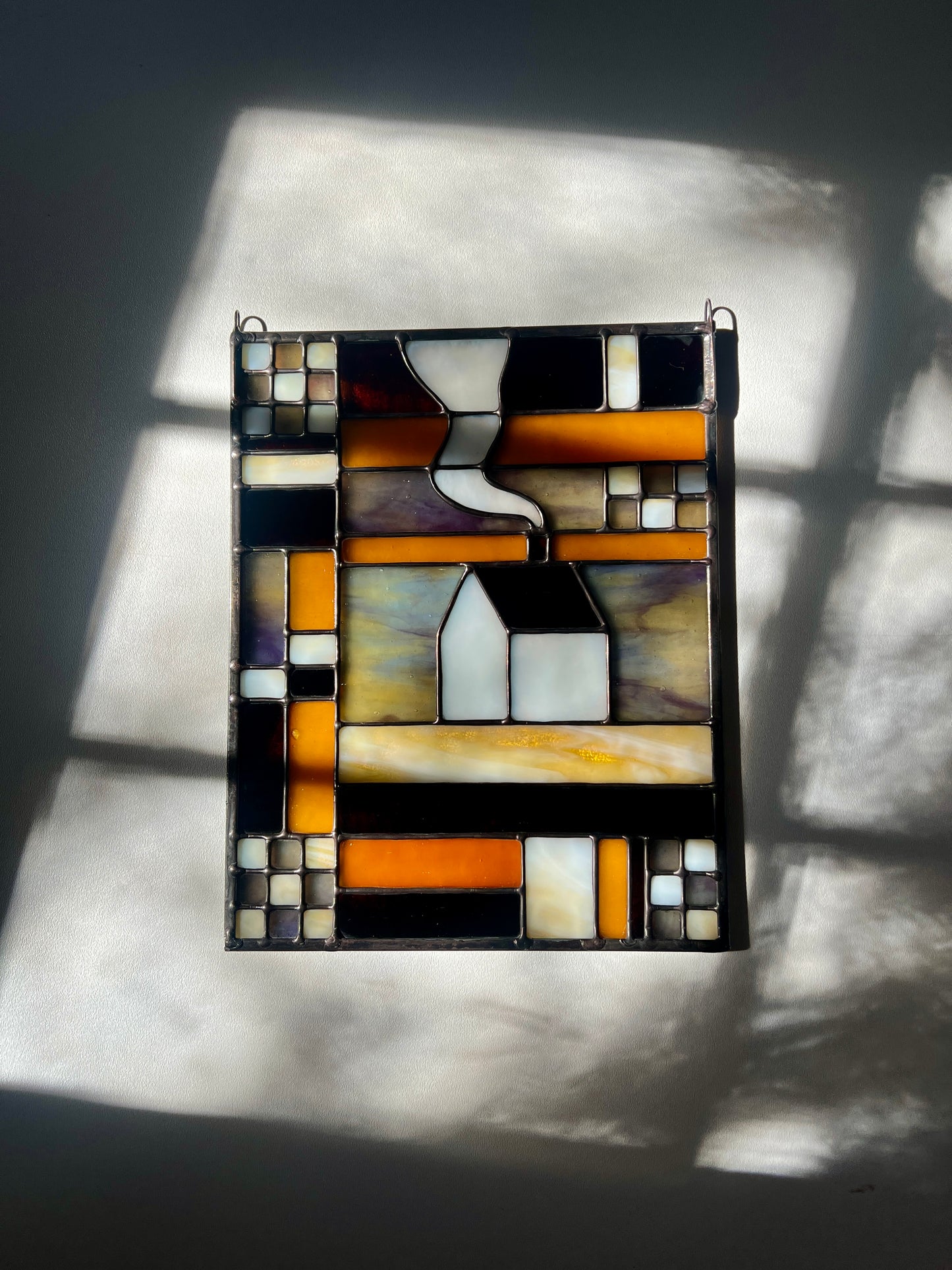 Stained glass textile inspired panel depicting a home in the center in shades of purple, white, brown, and amber, resting on a white surface with bright sunlight and shadows cast across
