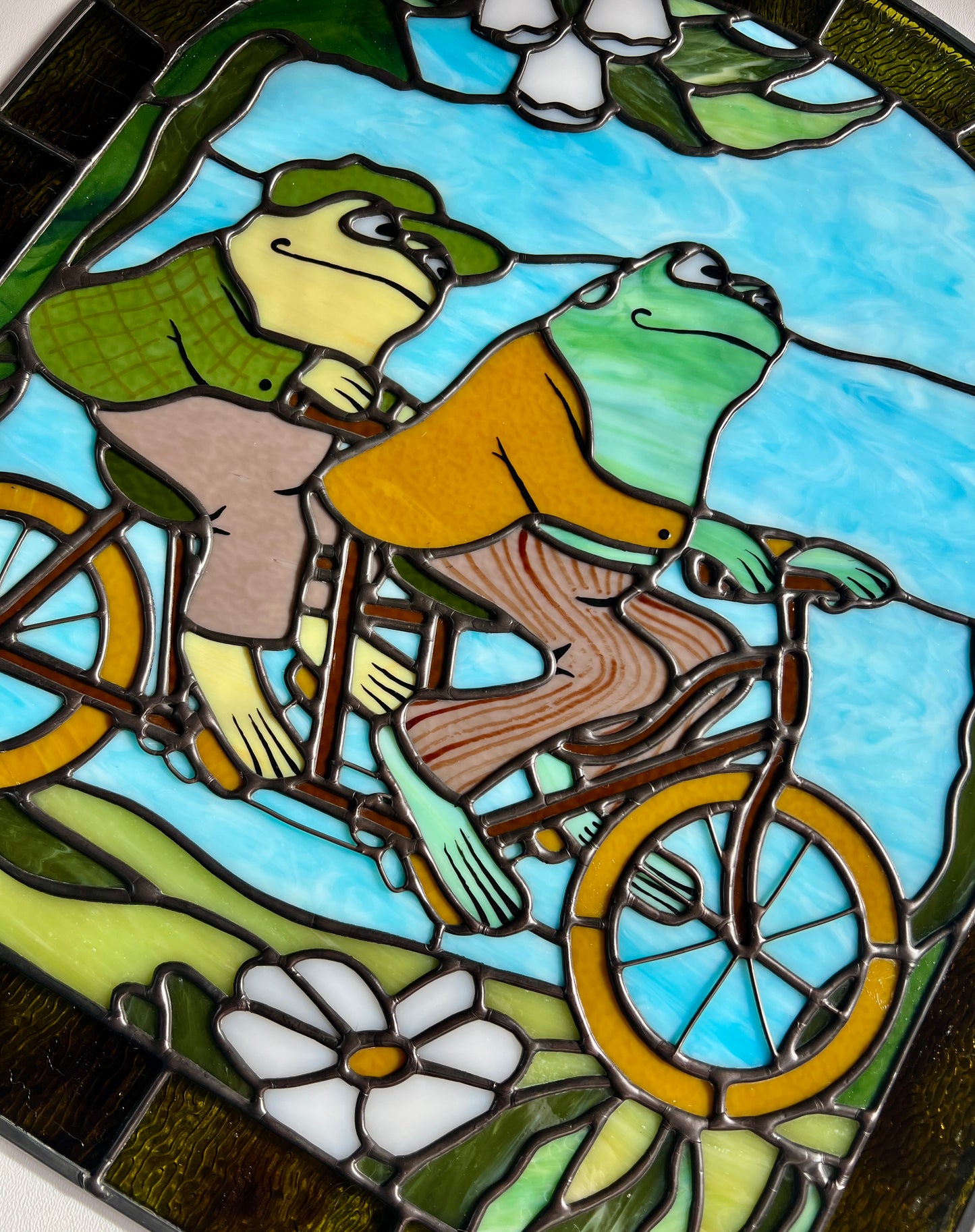 close up of a stained glass panel depicting frog and toad riding a bicycle resting on a white surface