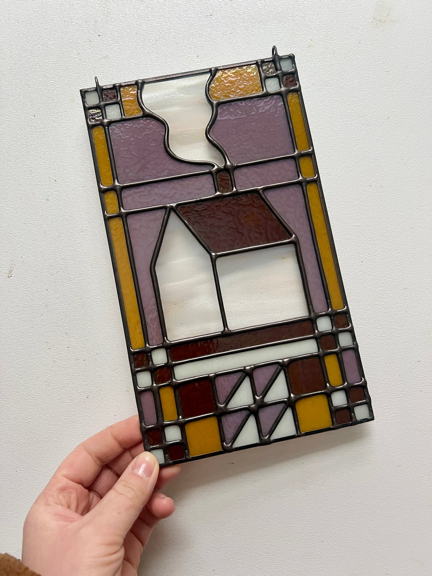 Stained glass textile inspired panel depicting a home in the center in shades of purple, white, brown, and bronze, held by a hand against a white surface