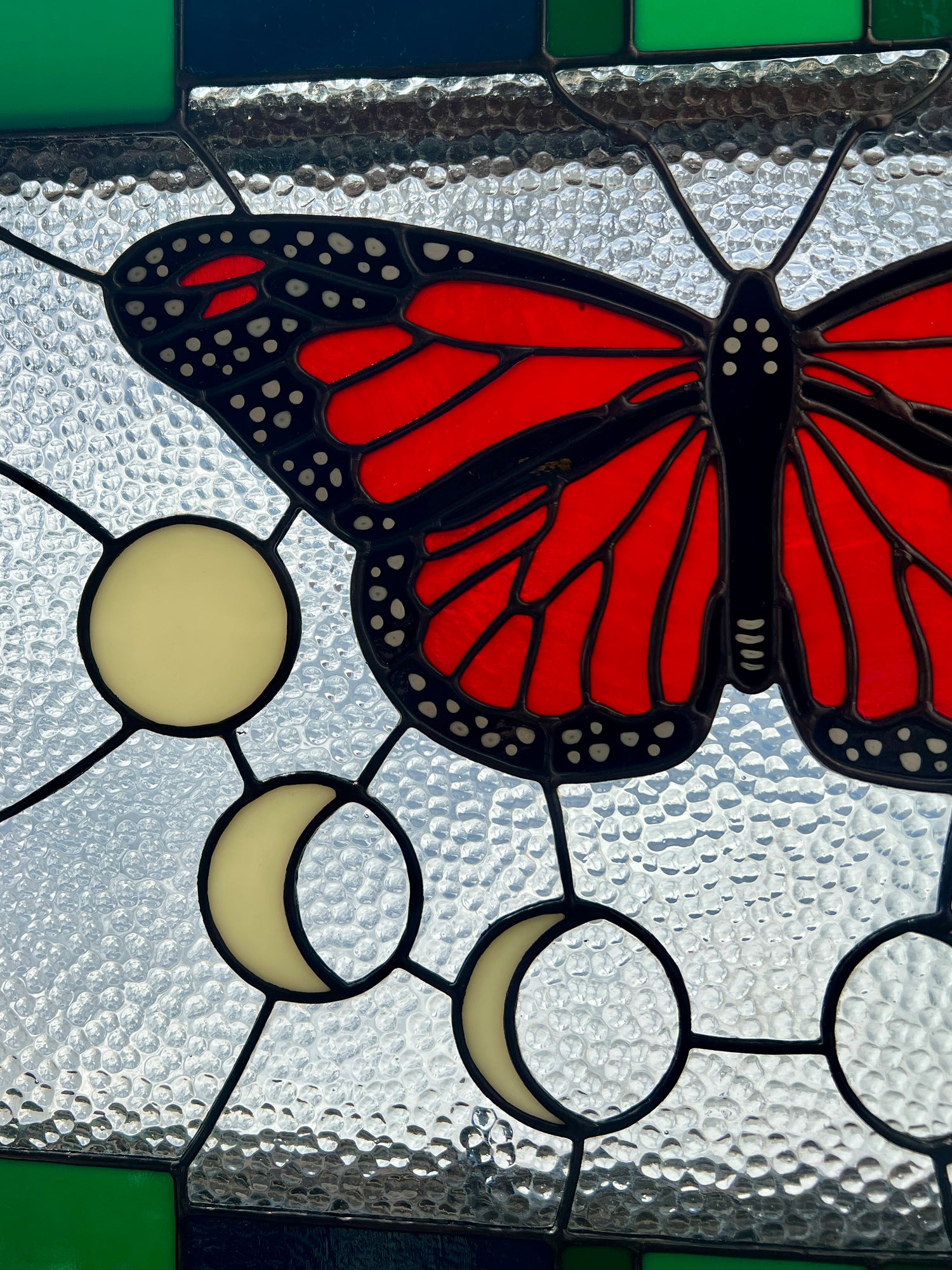 Close up of a stained glass panel with a thick blue and green geometric border and a monarch butterfly and moon phases in the center, with textured clear glass in the background. The panel is backlight demonstrating the bright glow of the reddish orange glass used for the butterfly.