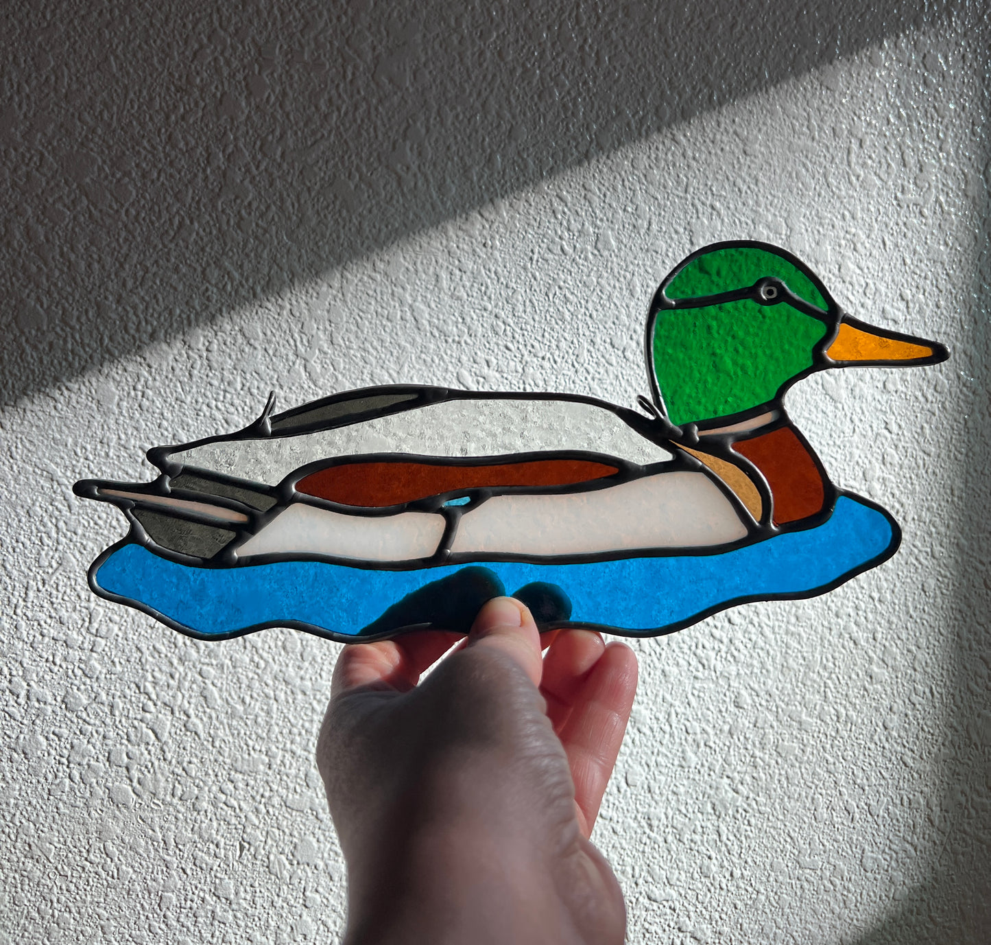 stained glass male mallard suncatcher held by a hand in front of a white wall