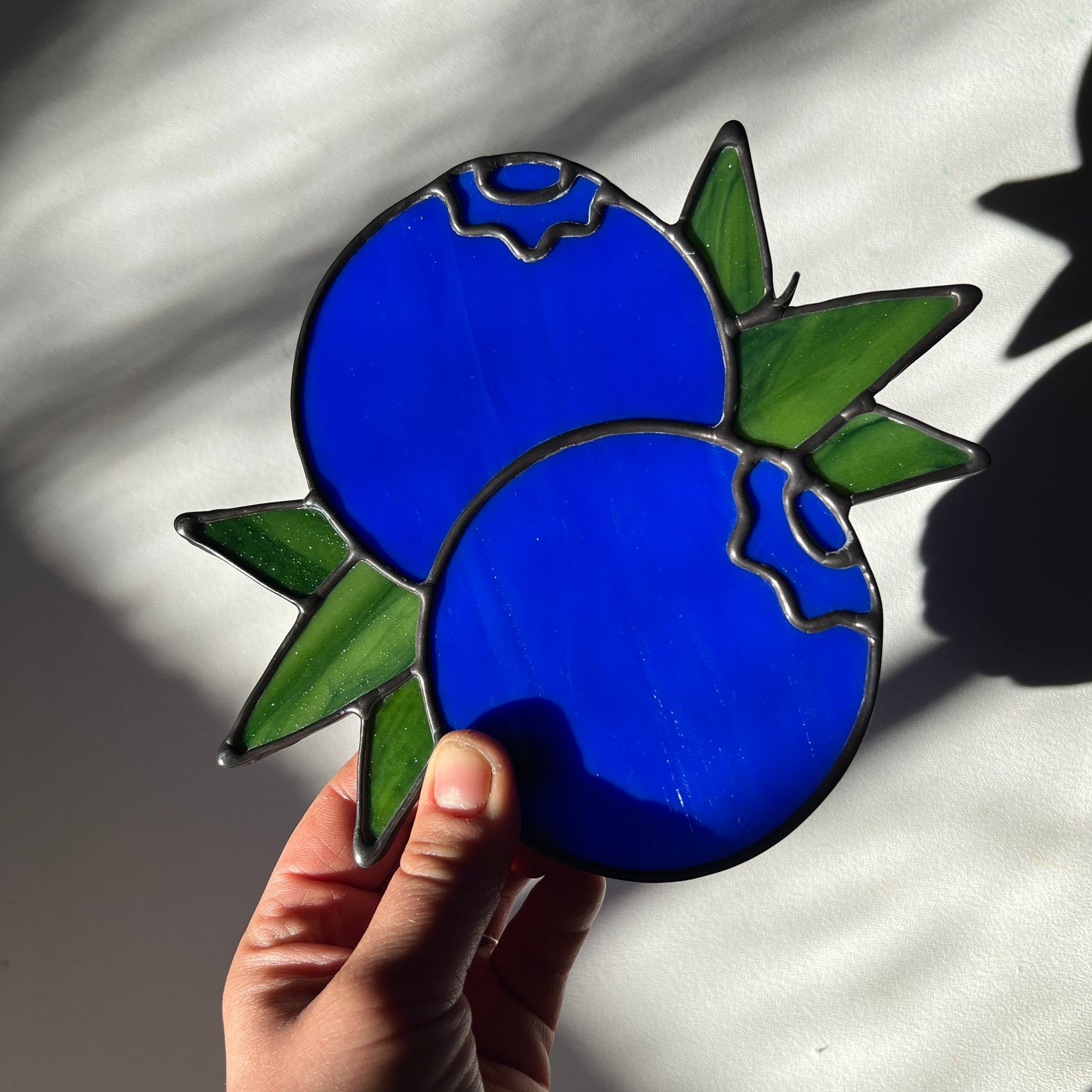 hand holding a stained glass suncatcher depicting a small bunch of blueberries with leaves