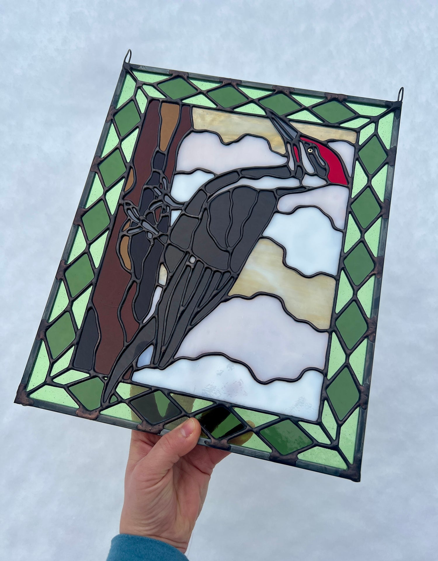 stained glass panel depicting a pileated woodpecker with a green diamond pattern border, held in someone's hand against a white background