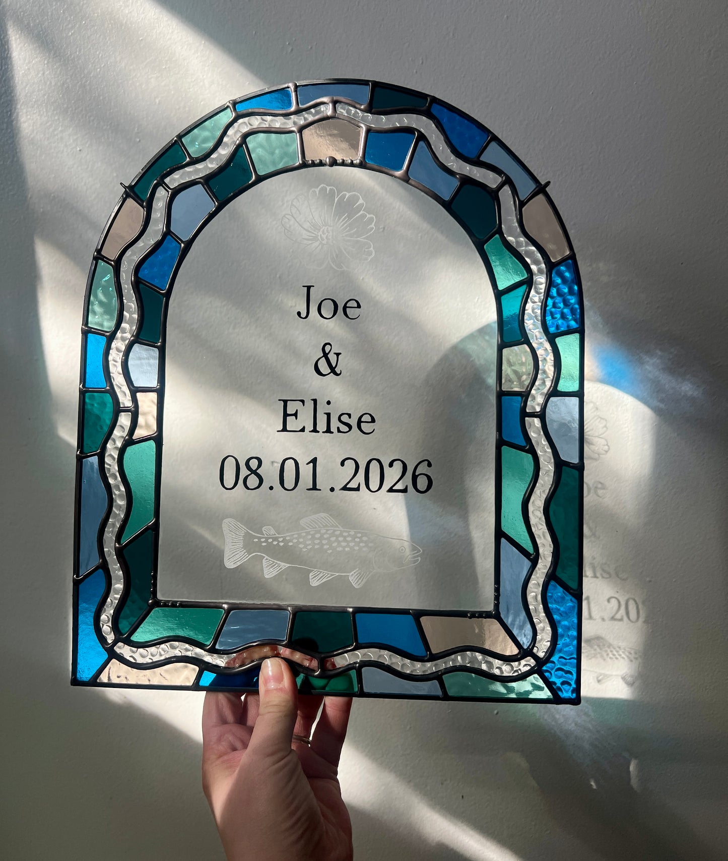 Stained glass panel in the shape of an arch with a multicolor blue, teal, turquoise, and pink border. Within the border is squiggle detail in clear textured glass. The center of the arch is clear uncolored glass, with personalized text and hand etched details of a cosmos flower and trout. The piece is held by a hand against a white wall, with gentle sunlight casting intermittent shadows and refractions.