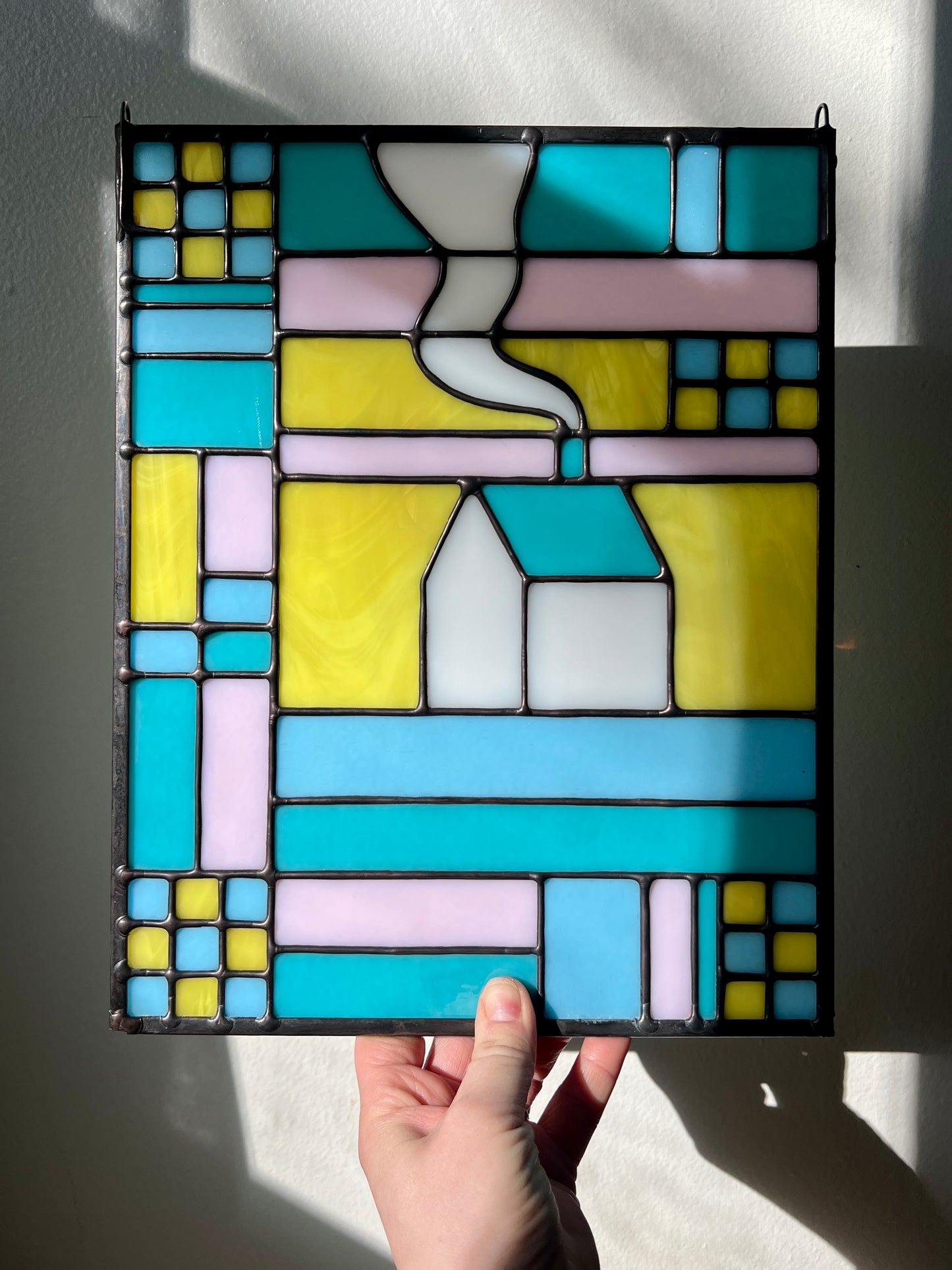 Stained glass textile inspired panel depicting a home in the center in shades of teal, pink, white, blue, and yellow, held by a hand in front of a white background