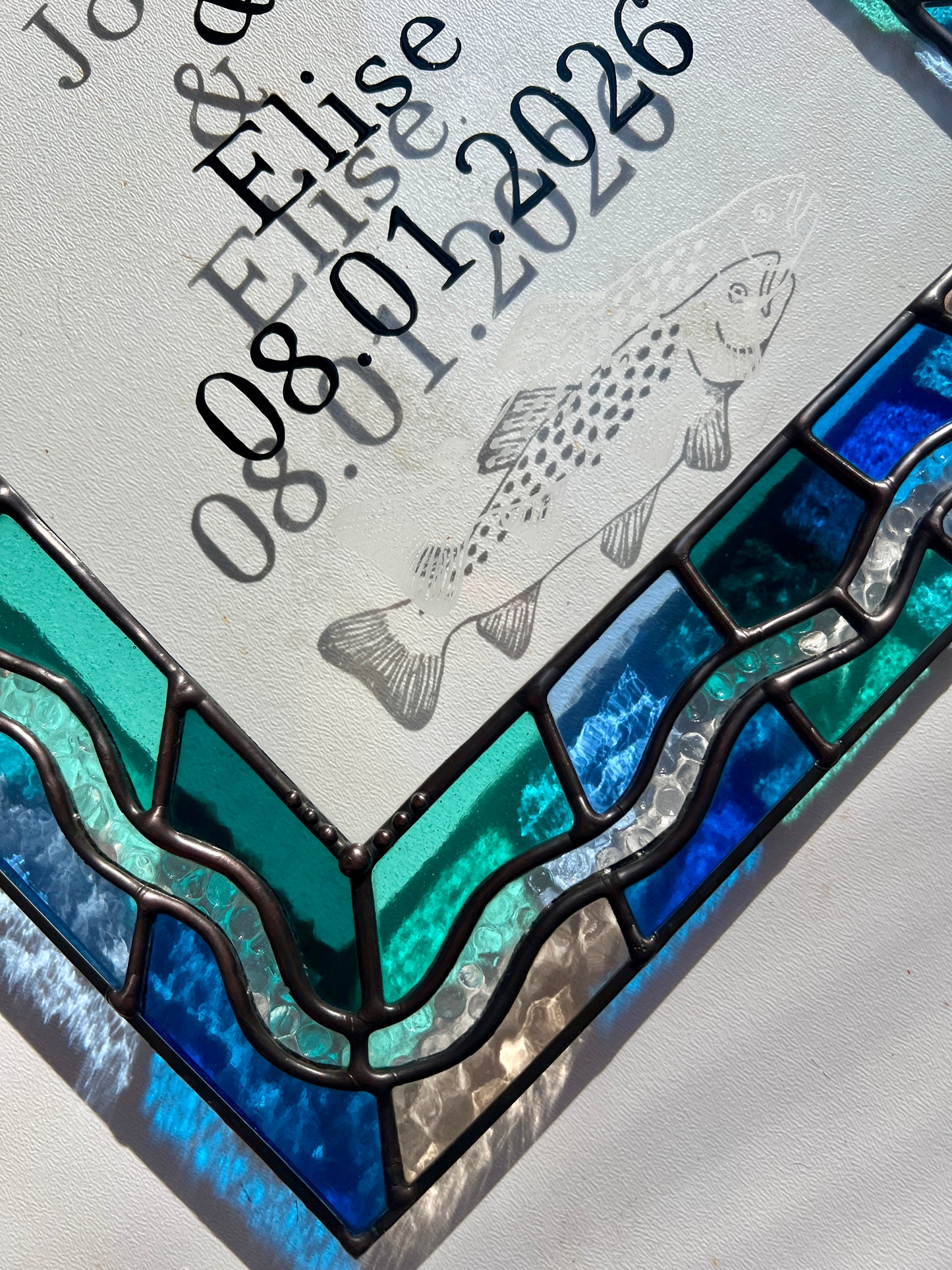 Close up of a stained glass panel in the shape of an arch with a multicolor blue, teal, turquoise, and pink border. Within the border is squiggle detail in clear textured glass. The center of the arch is clear uncolored glass, with personalized text and hand etched details of a trout. Sunlight casts refractions and shadows of the text, colored glass, and etchings.