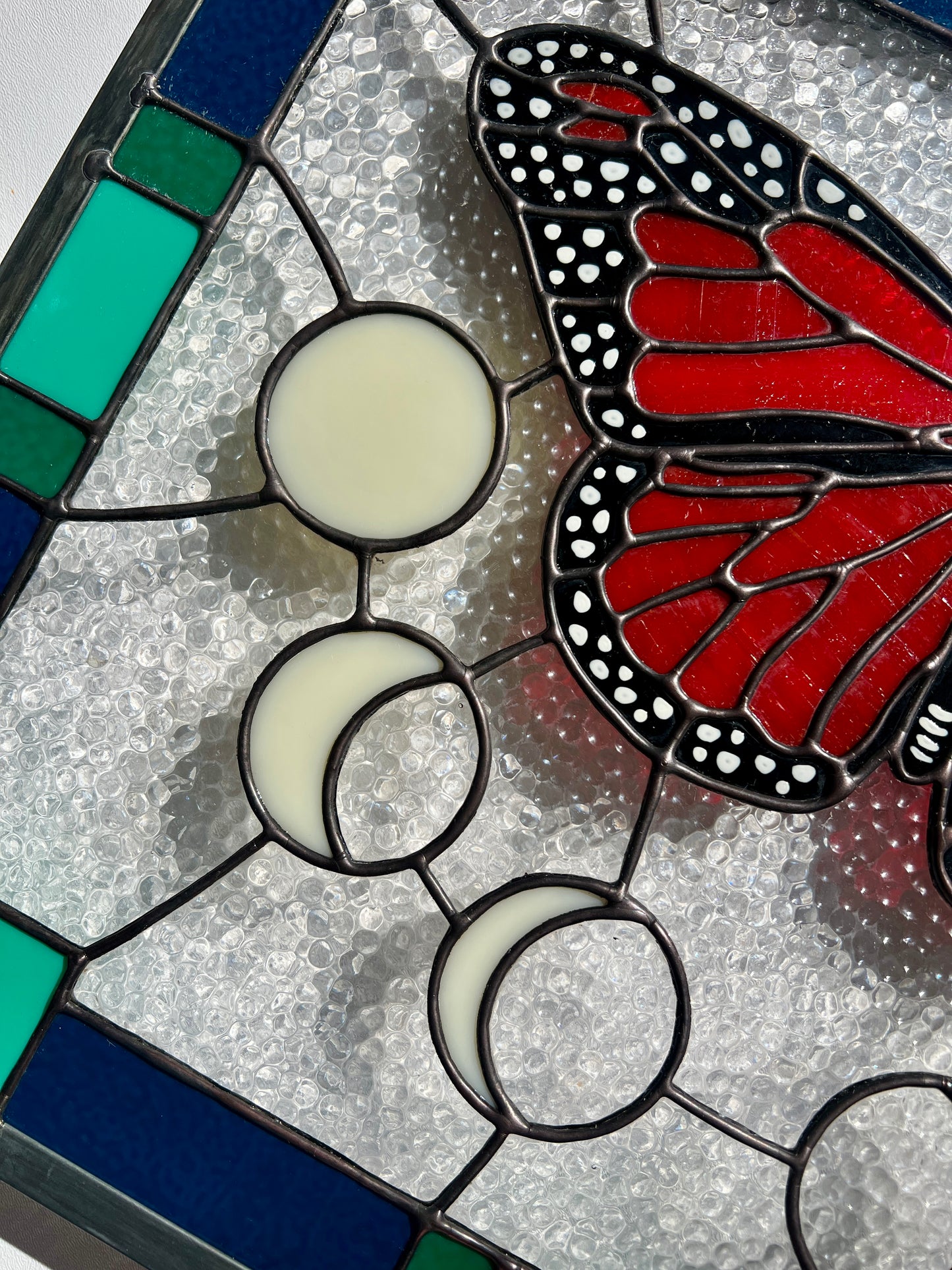 Close up of a large stained glass panel with a thick blue and green geometric border and a monarch butterfly and moon phases in the center, with textured clear glass in the background.
