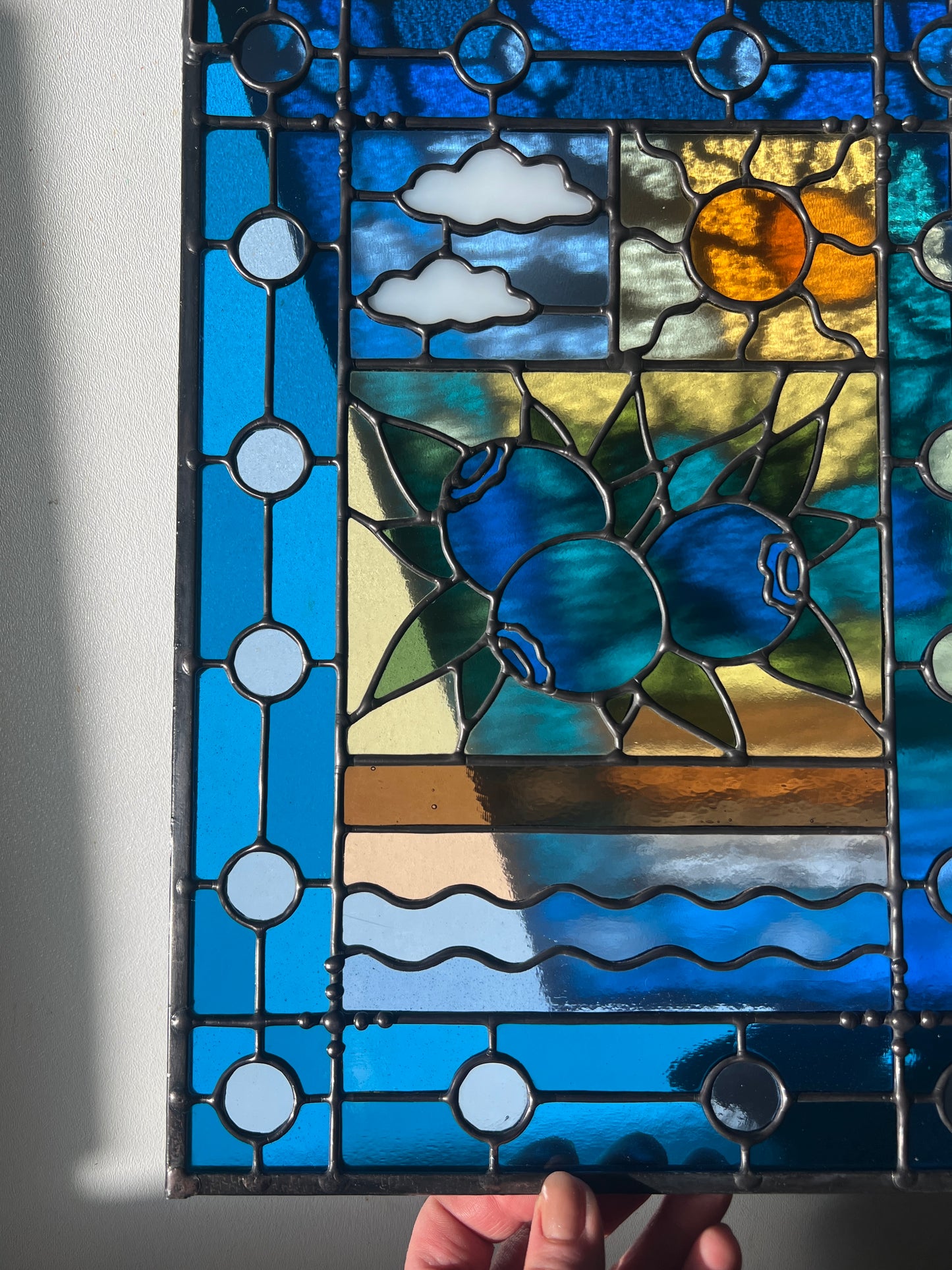 close up of stained glass panel with a simple compartment style design featuring clouds, sun, water, and a small bunch of blueberries in the middle with a blue border, with sunlight passing through and casting a colorful refraction