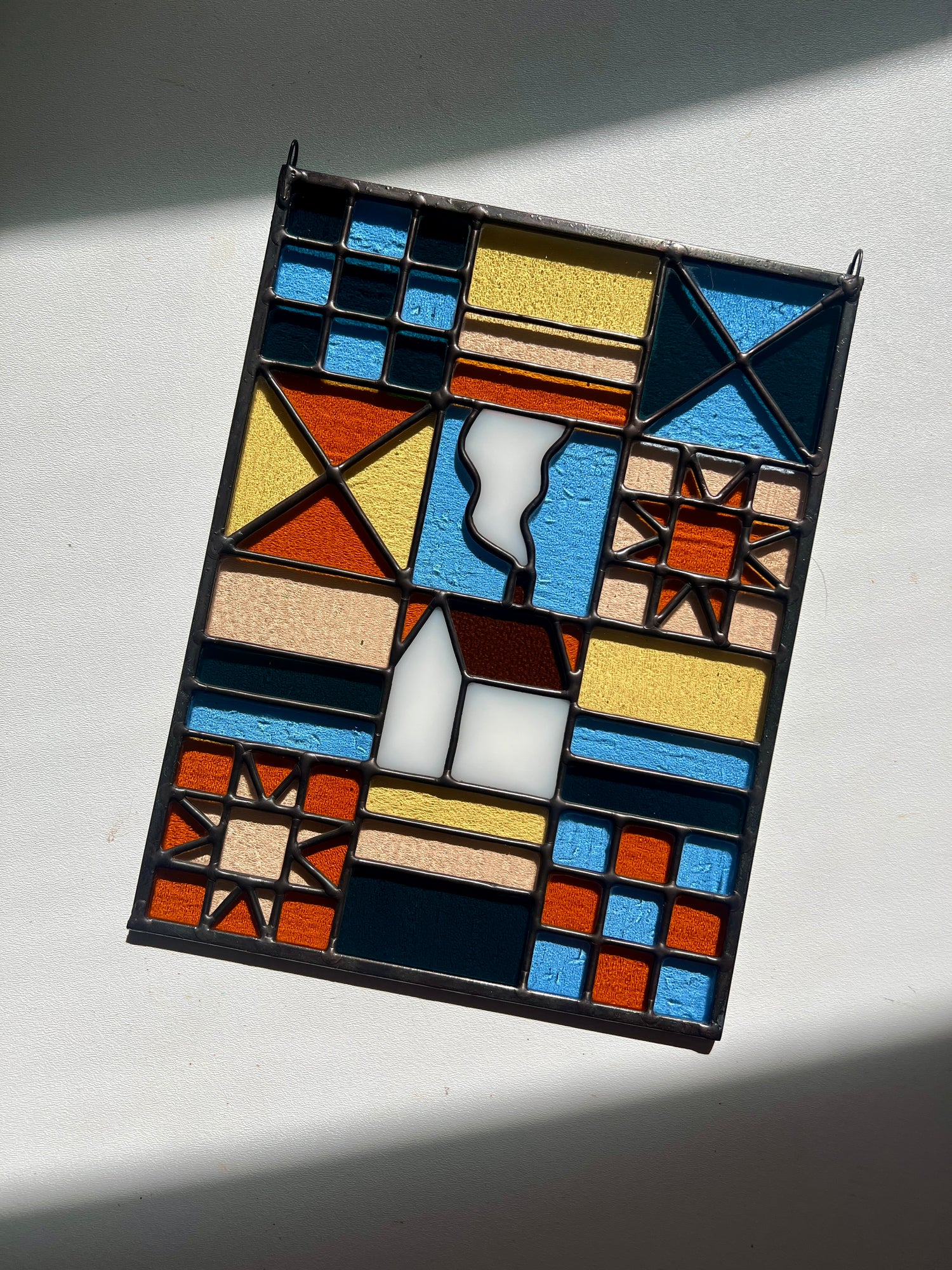 Stained glass textile inspired panel depicting a home in the center in shades of teal, pink, white, blue, and yellow, against a white background with bright sunlight across